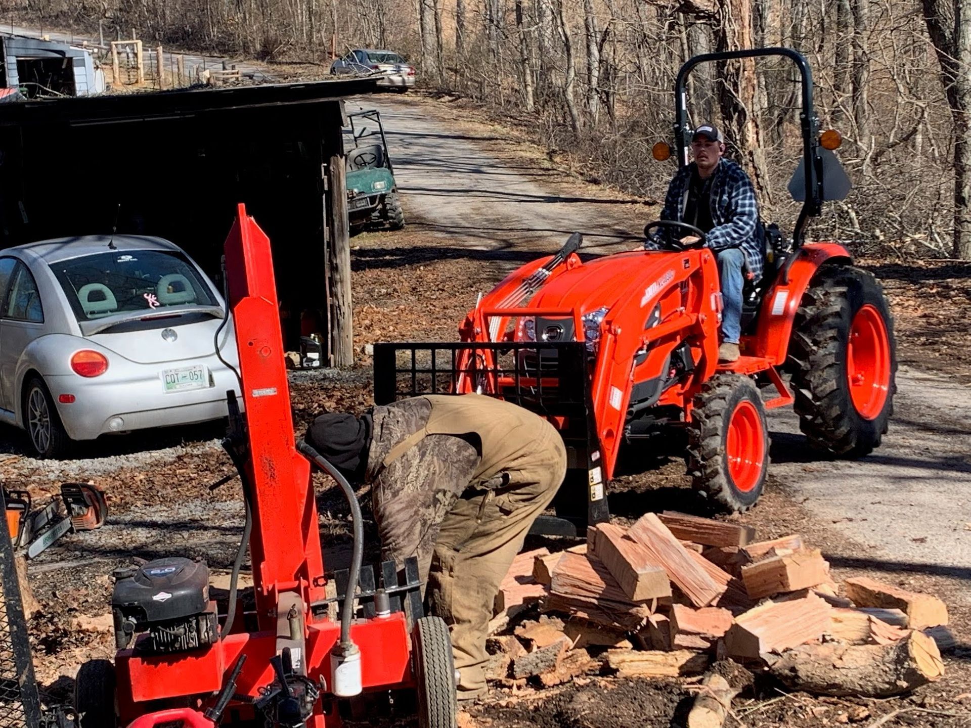 A person in camouflage gear sorts wood by a log splitter while another sits on a bright orange Kubota tractor outdoors.