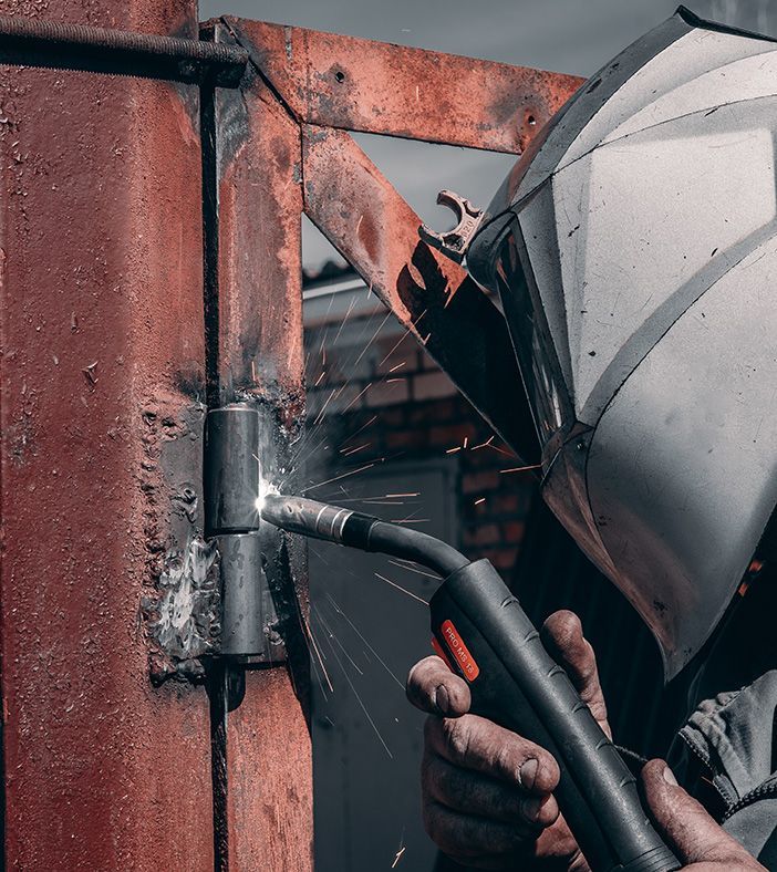 A person wearing a protective welding helmet uses a MIG torch to weld a metal hinge onto a rusty metal gate frame.