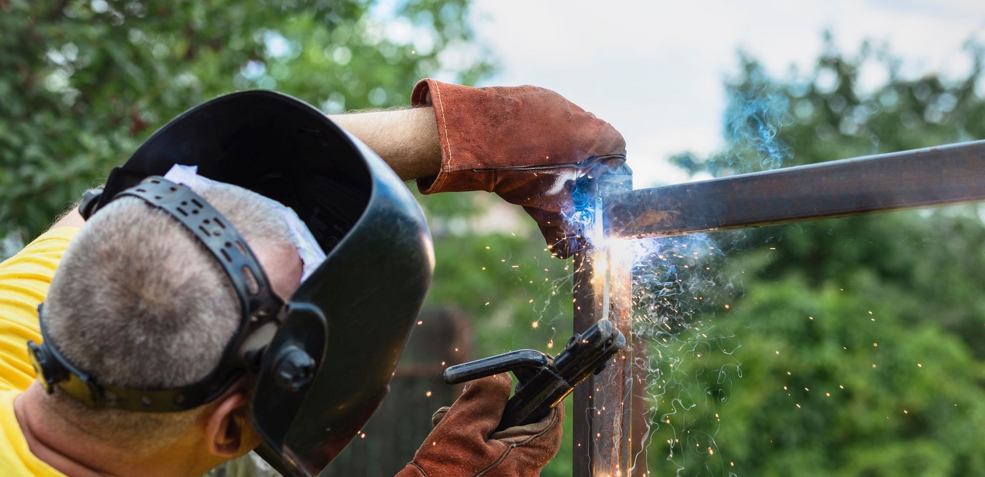 A person wearing a protective welding helmet and leather gloves uses a welding torch to join two pieces of metal outdoors.