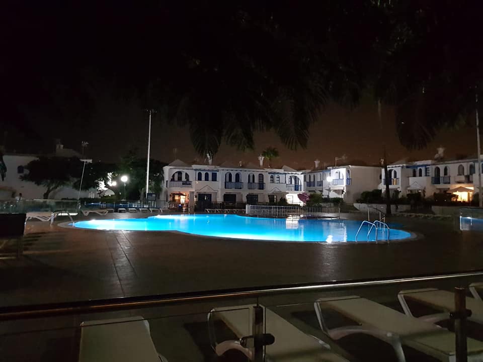 Vor einer Häuserzeile ist ein großes Schwimmbad, das nachts beleuchtet ist.