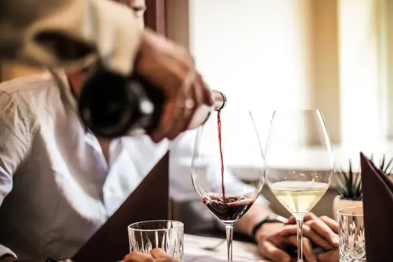Person pouring red wine into a glass at a restaurant table with other wine glasses and a person holding a camera