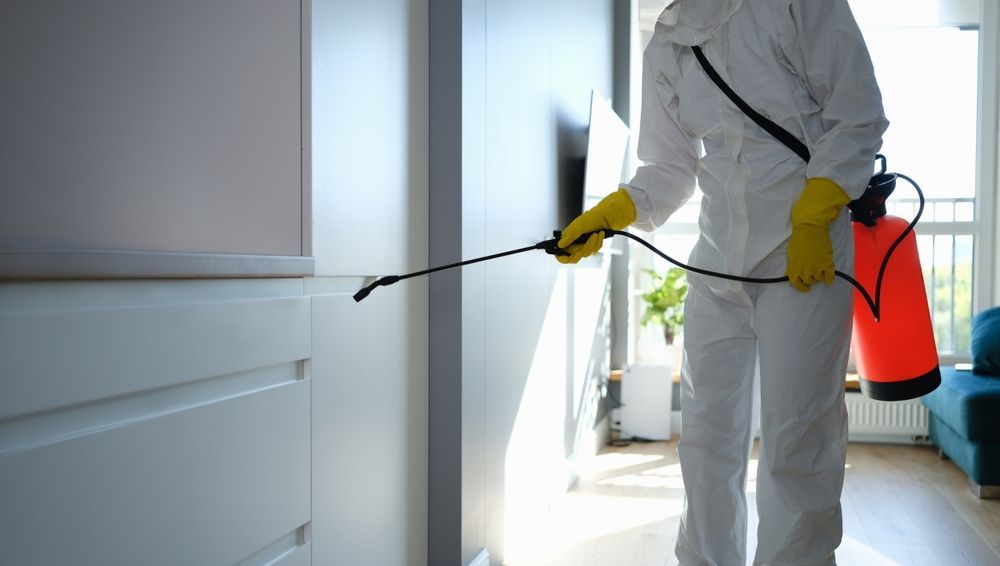 Person in protective suit spraying insecticide inside a room with a red tank.