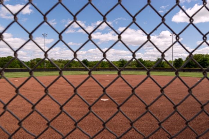 Softball field viewed through chain-link fence, under a partly cloudy blue sky.