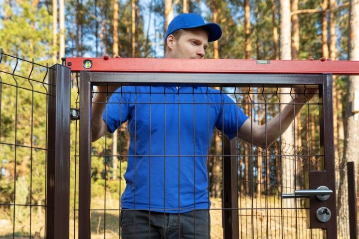 Man in blue shirt and cap uses a level to install a gate in a wire fence, outdoors.