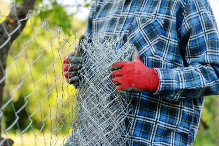 Person in plaid shirt and red gloves holding rolled-up chain-link fence outdoors.