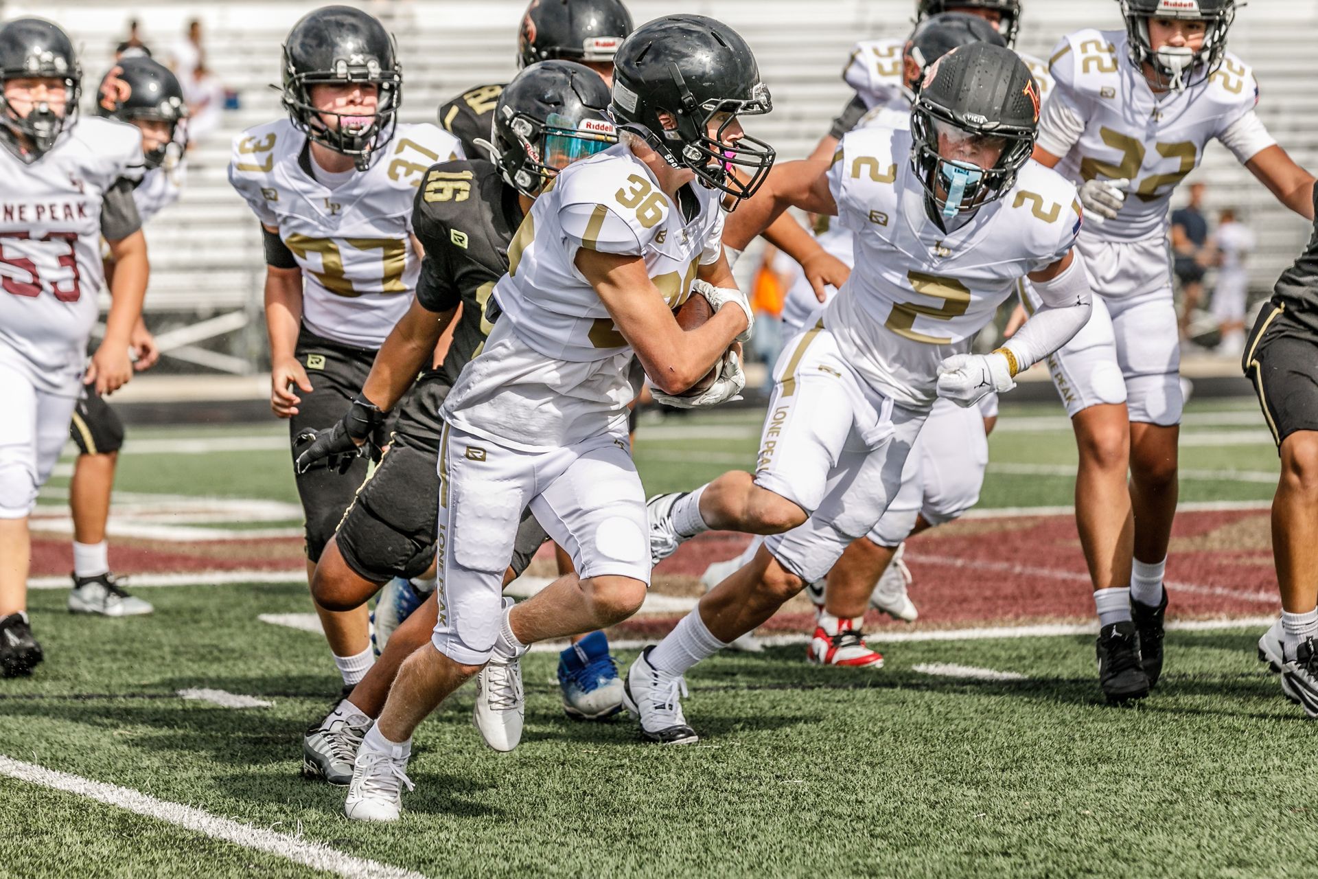 Football players in action on a grassy field; one runs with the ball, pursued by two opponents.