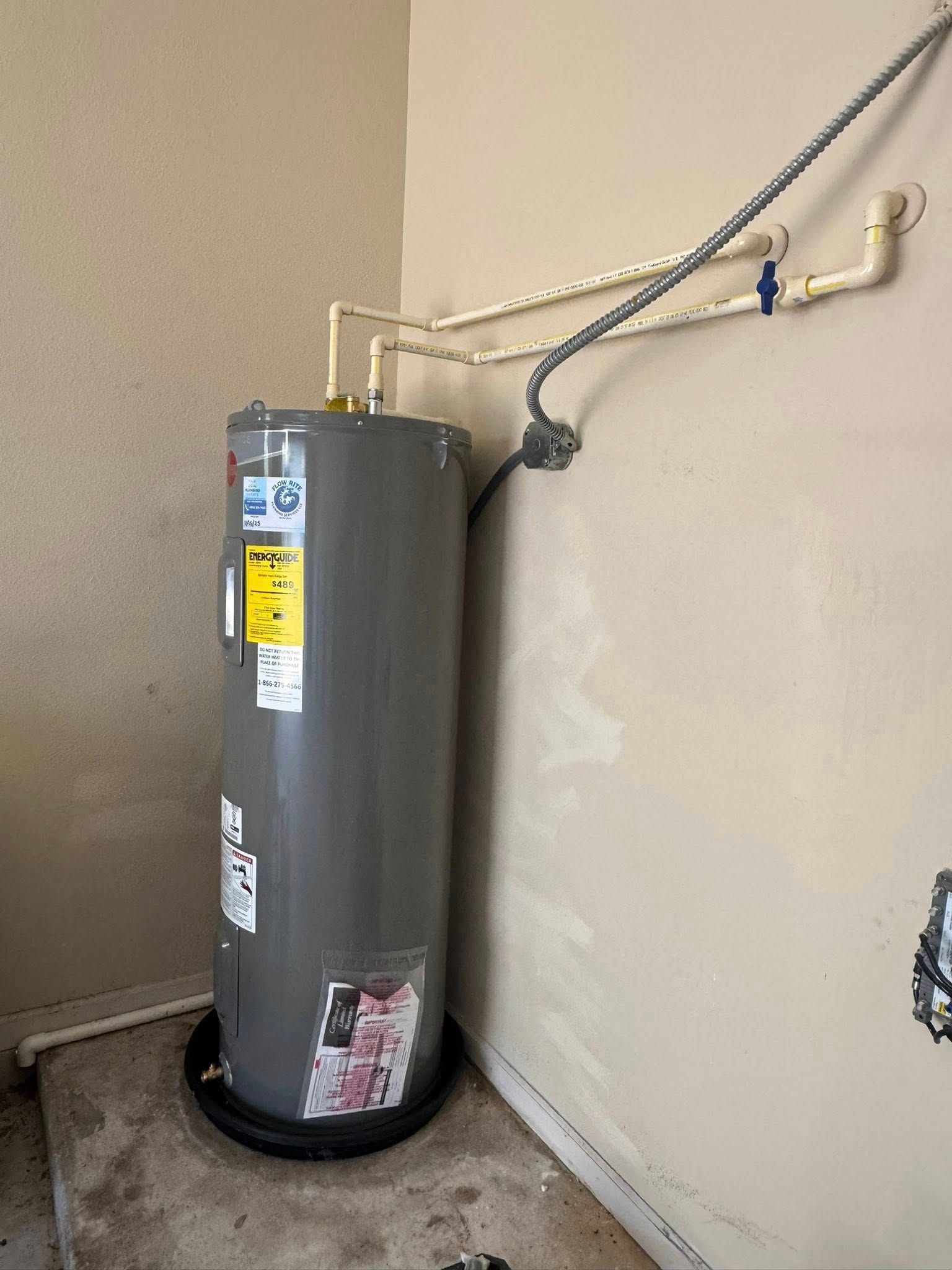 A gray electric water heater sits in a drip pan on a concrete floor with exposed plumbing pipes running along a wall.