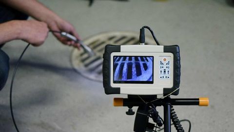 A device with a video monitor showing a pipe interior, positioned on a tripod near a person holding a sewer inspection probe.