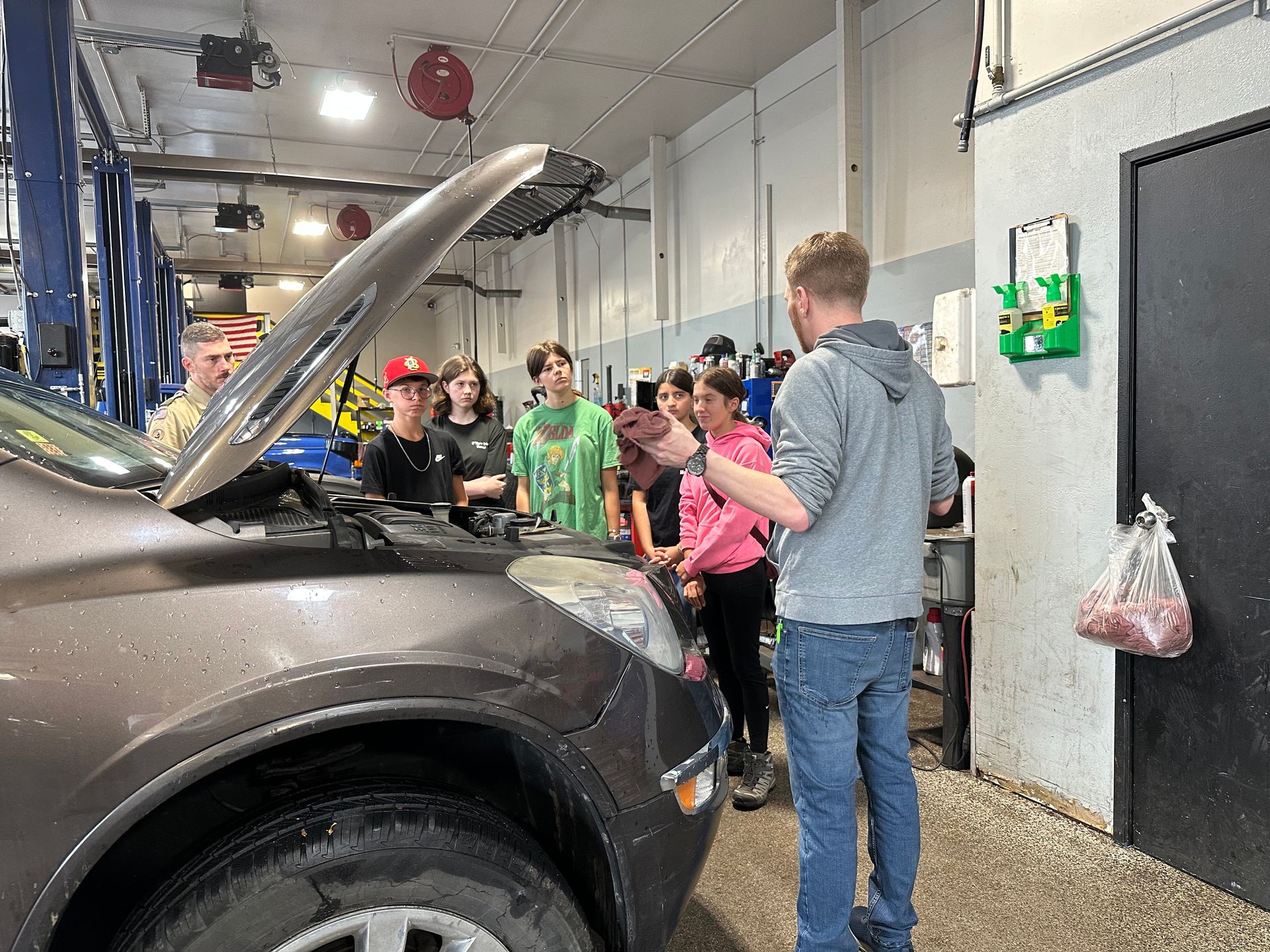 A group of students in a garage listen to an instructor as he points at a car engine with the hood open.