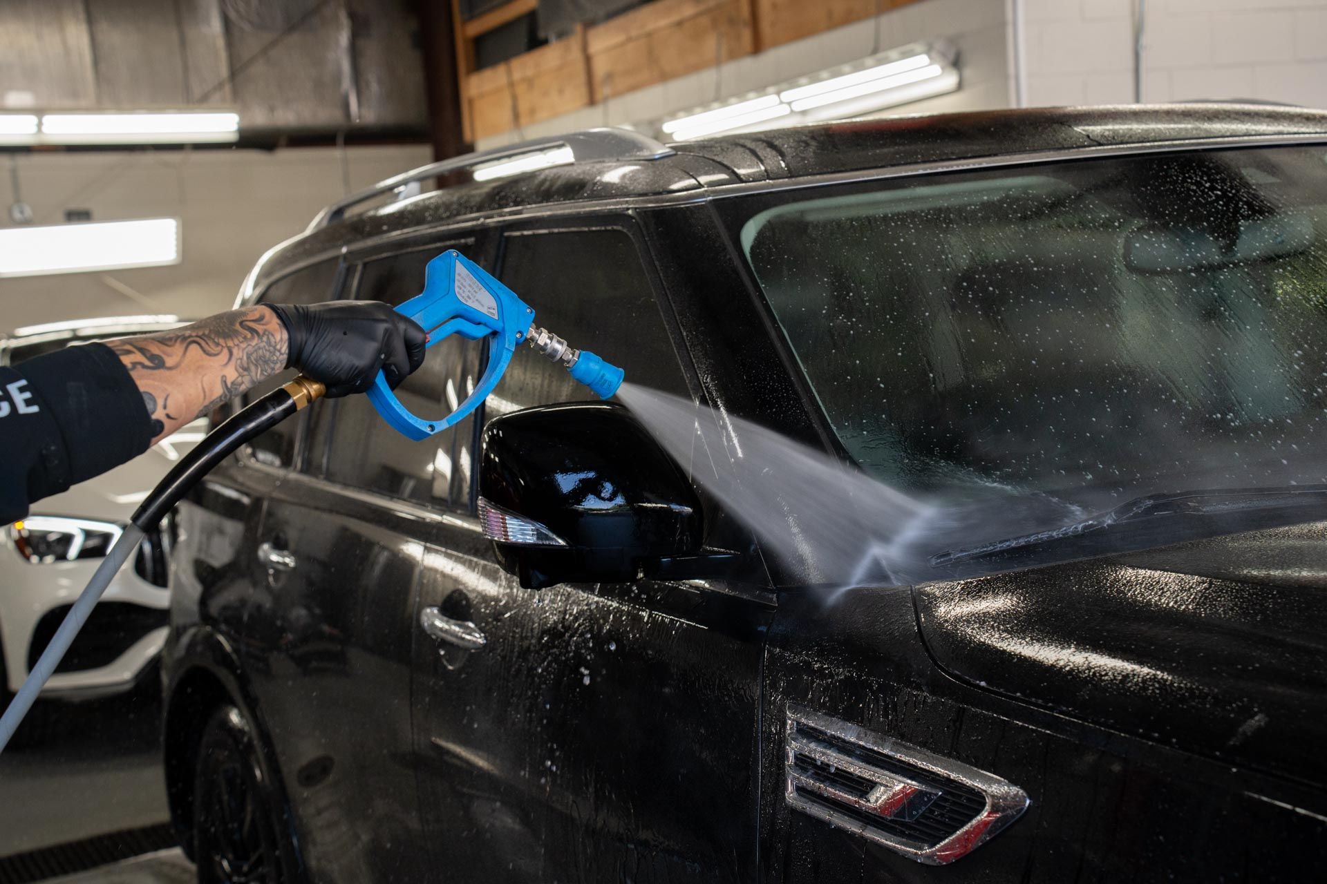Person in black gloves washing a black SUV with a blue spray nozzle in a garage.
