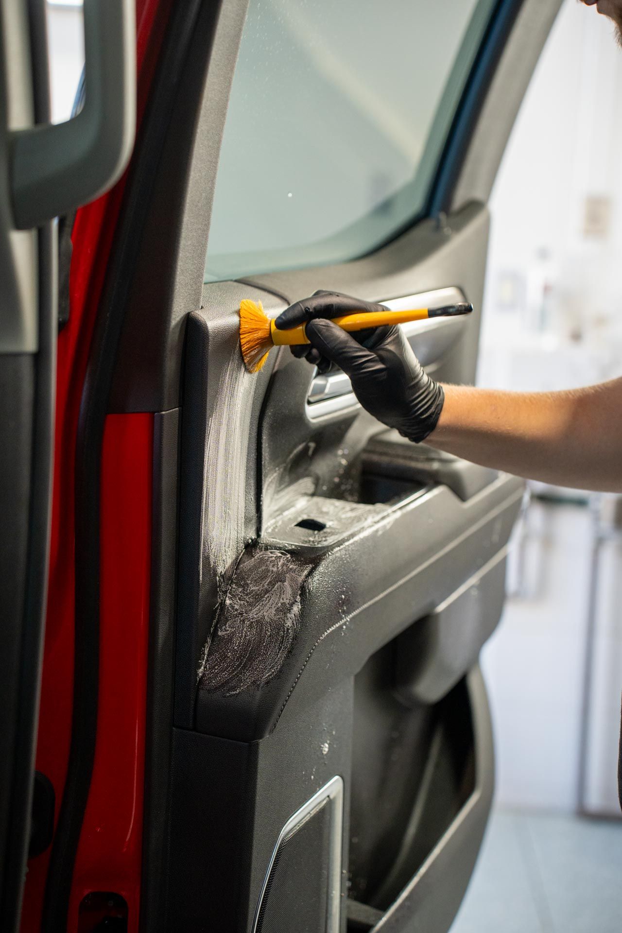 Person wearing a black glove brushes interior car panel. Red car door, black trim, yellow brush.