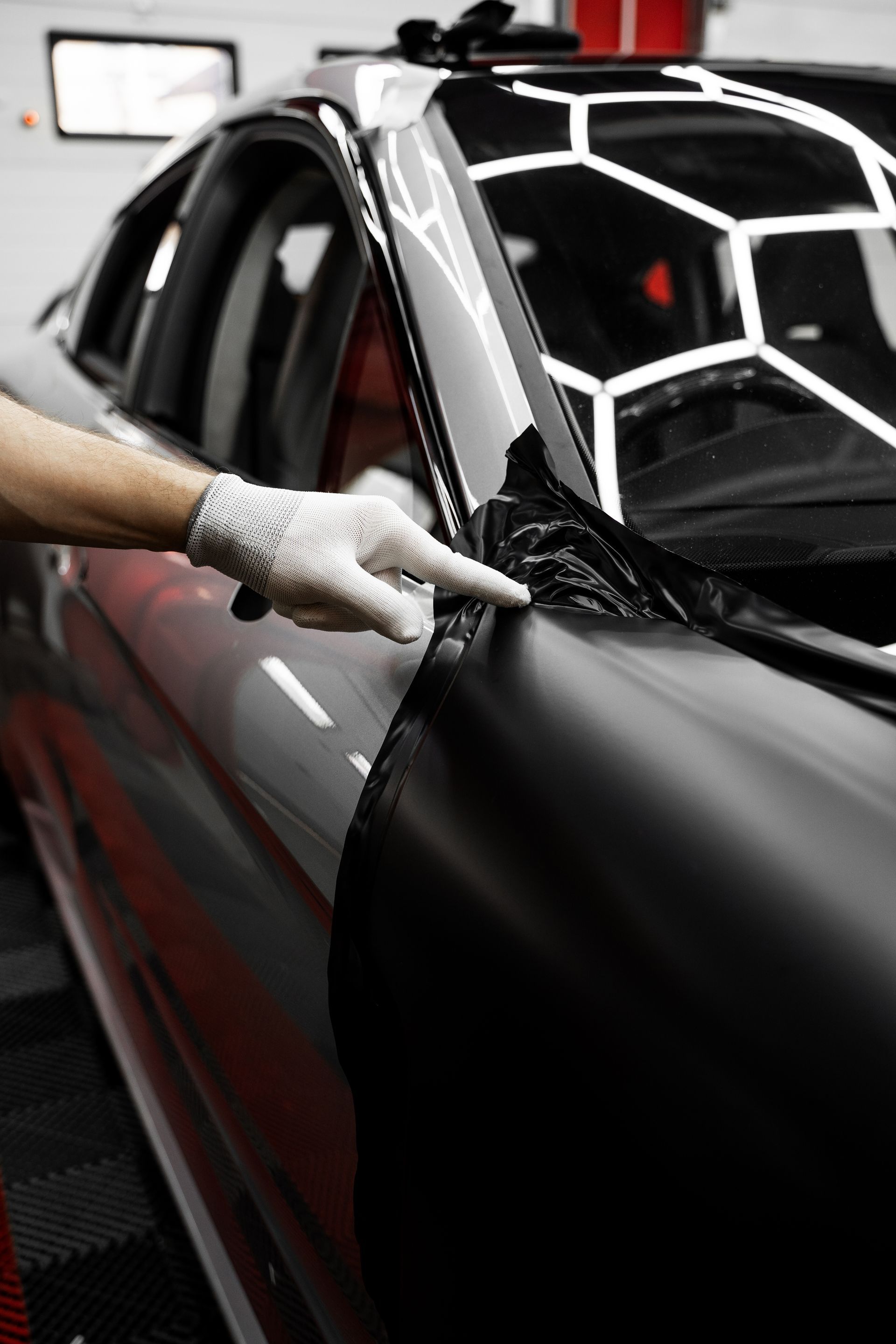 Person wearing a glove points at black vinyl on a car during application, in a garage setting.