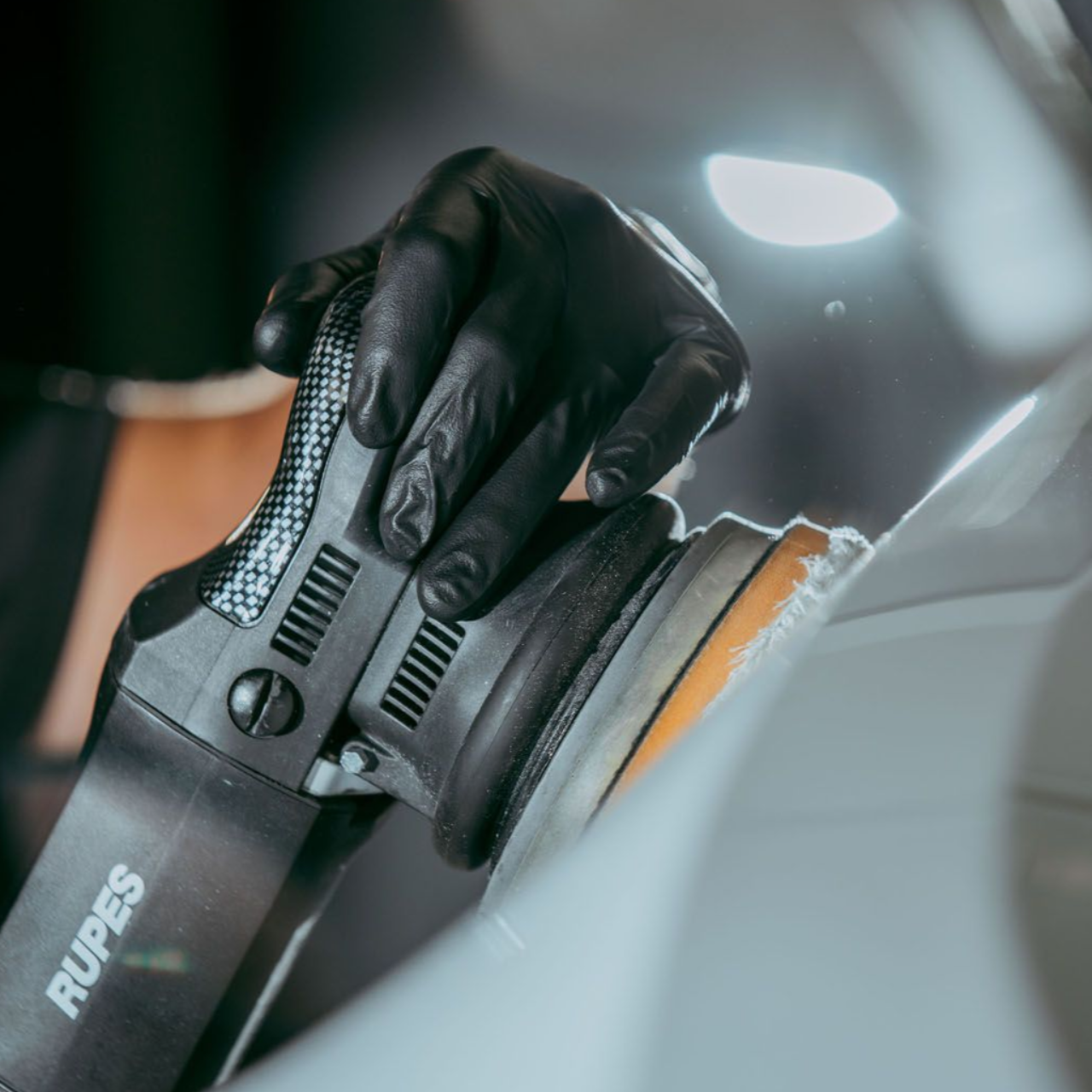 Gloved hand using a Rupes polisher on a white car surface, detail work.