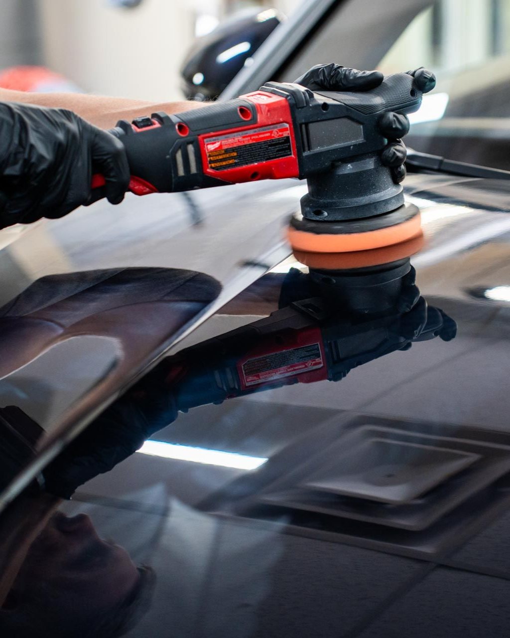 A gloved hand polishes a car's black surface with a red and black power buffer.