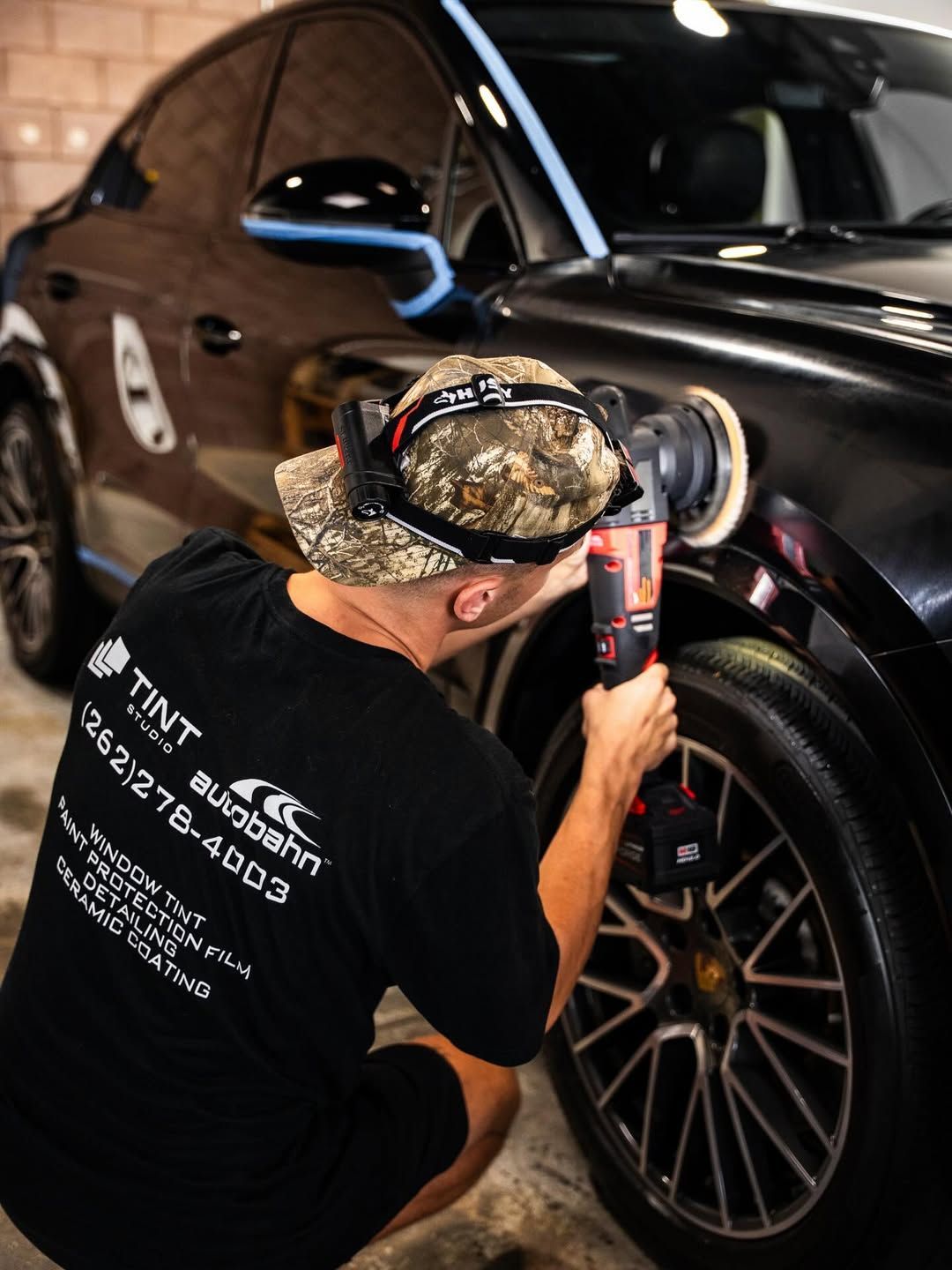 Person polishing a black car with a rotary buffer, outdoors.