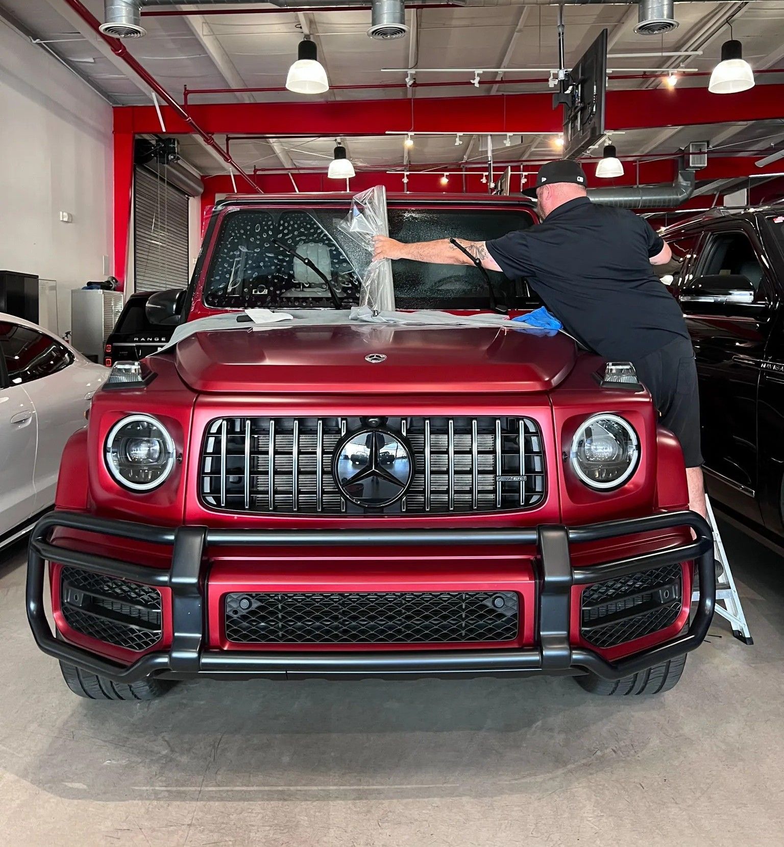 Man applying film to the windshield of a red Mercedes-Benz G-Wagon in a shop.