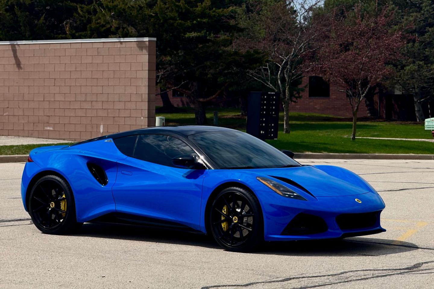 Blue sports car with black wheels and yellow brake calipers parked near a brick building and trees.