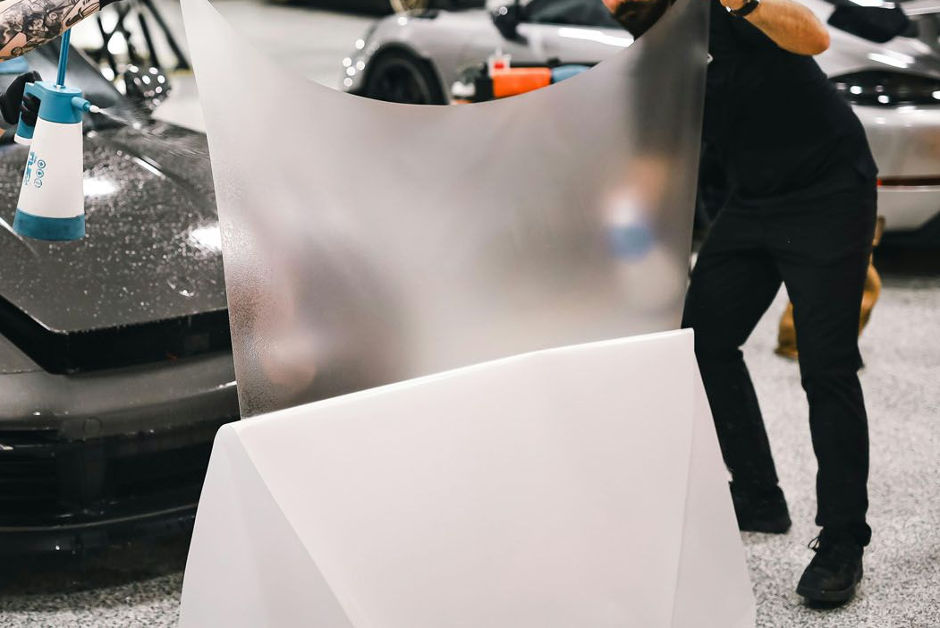 Two people applying film to a grey car in a workshop. One sprays water. Cars in background.