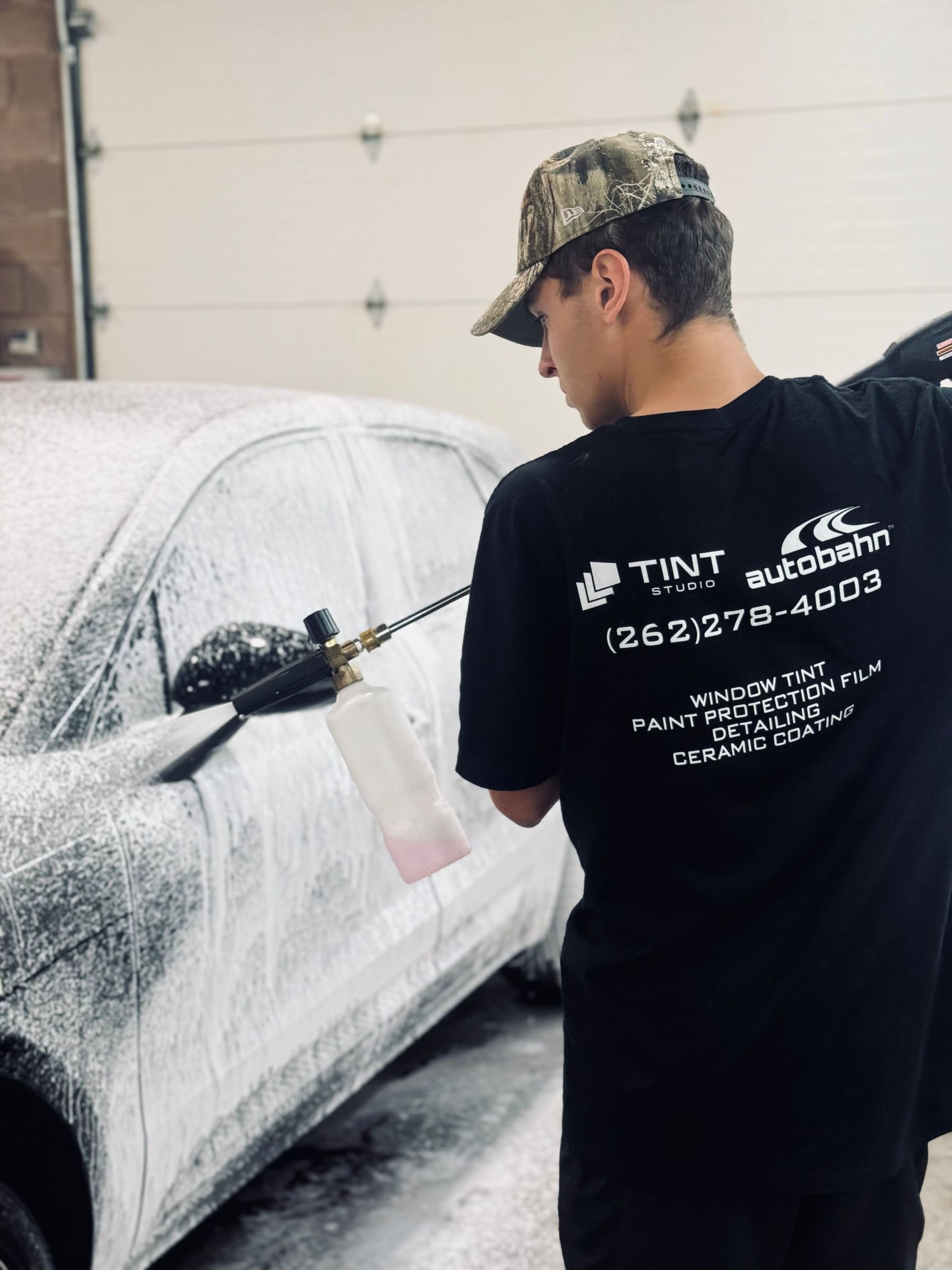 Person washing a car with foam in a garage, wearing a black shirt and camouflage hat.
