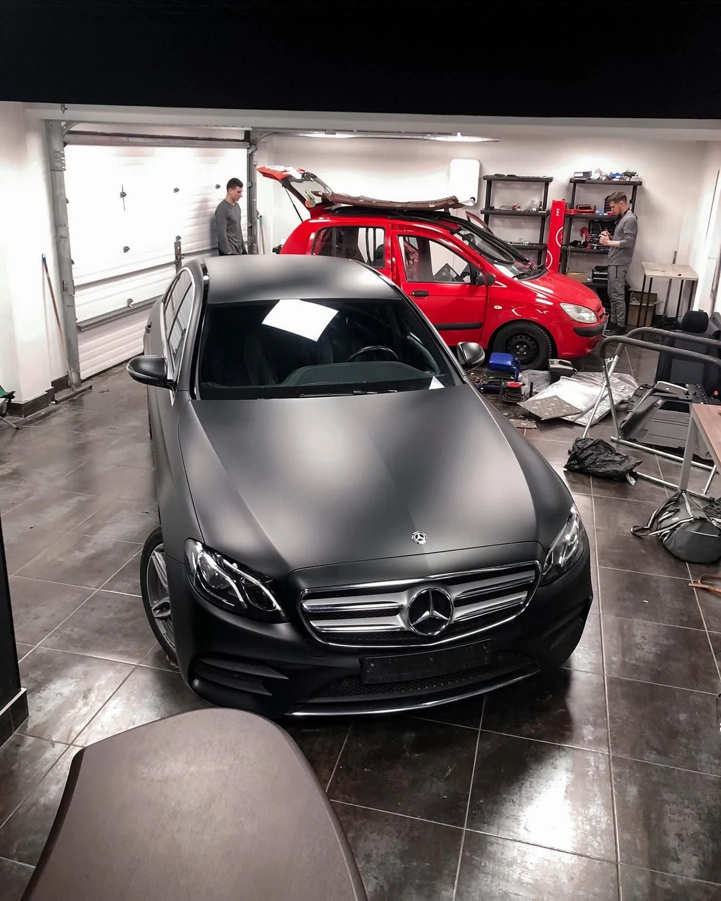 Black matte Mercedes sedan in a garage, red SUV in the background, two people working.