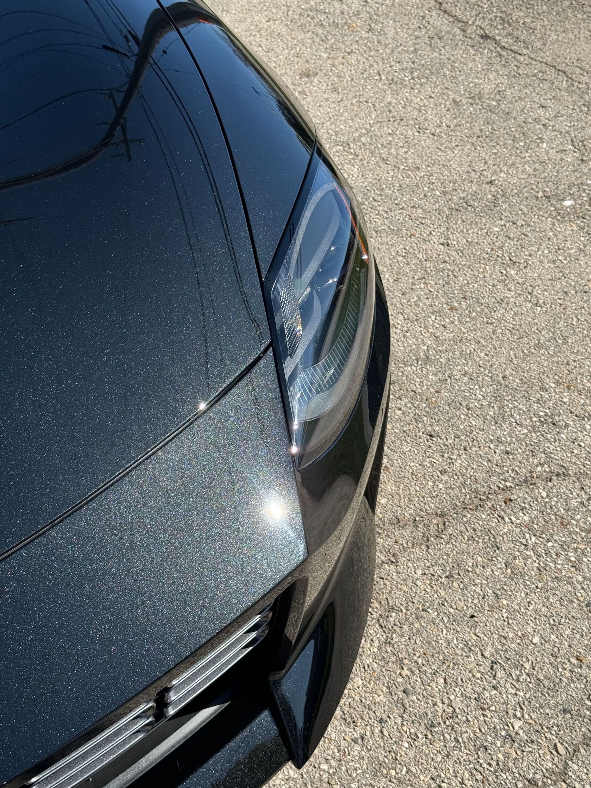 Black car front end, showing headlight, hood with sparkly finish, and part of the grill on a paved surface.