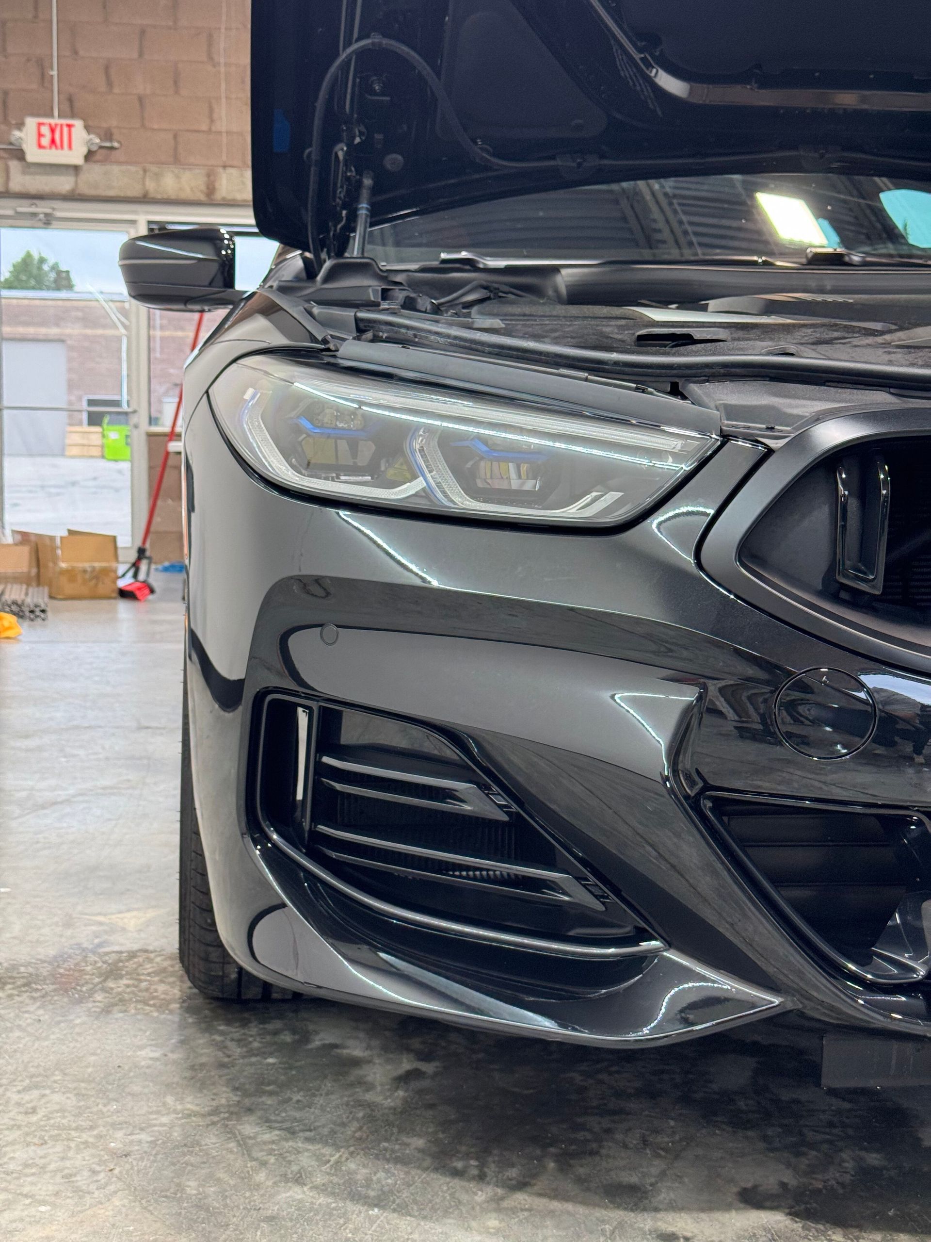 Black car, hood open, front corner view. In a garage setting with bright lights.