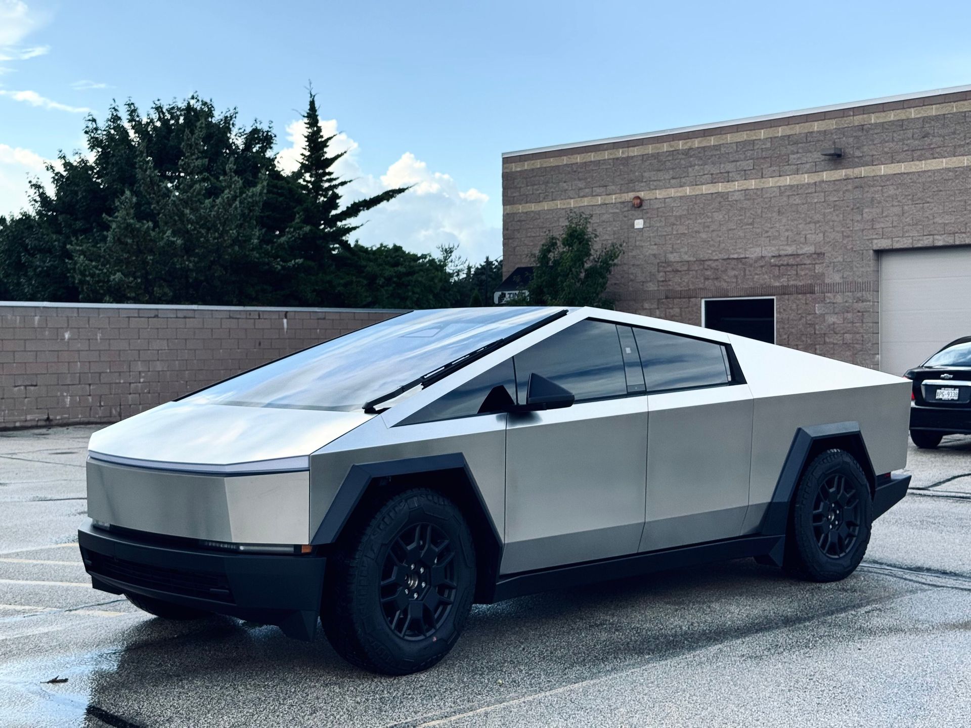 Tesla Cybertruck, silver, parked on wet pavement in front of a brick building and a tree-lined area.