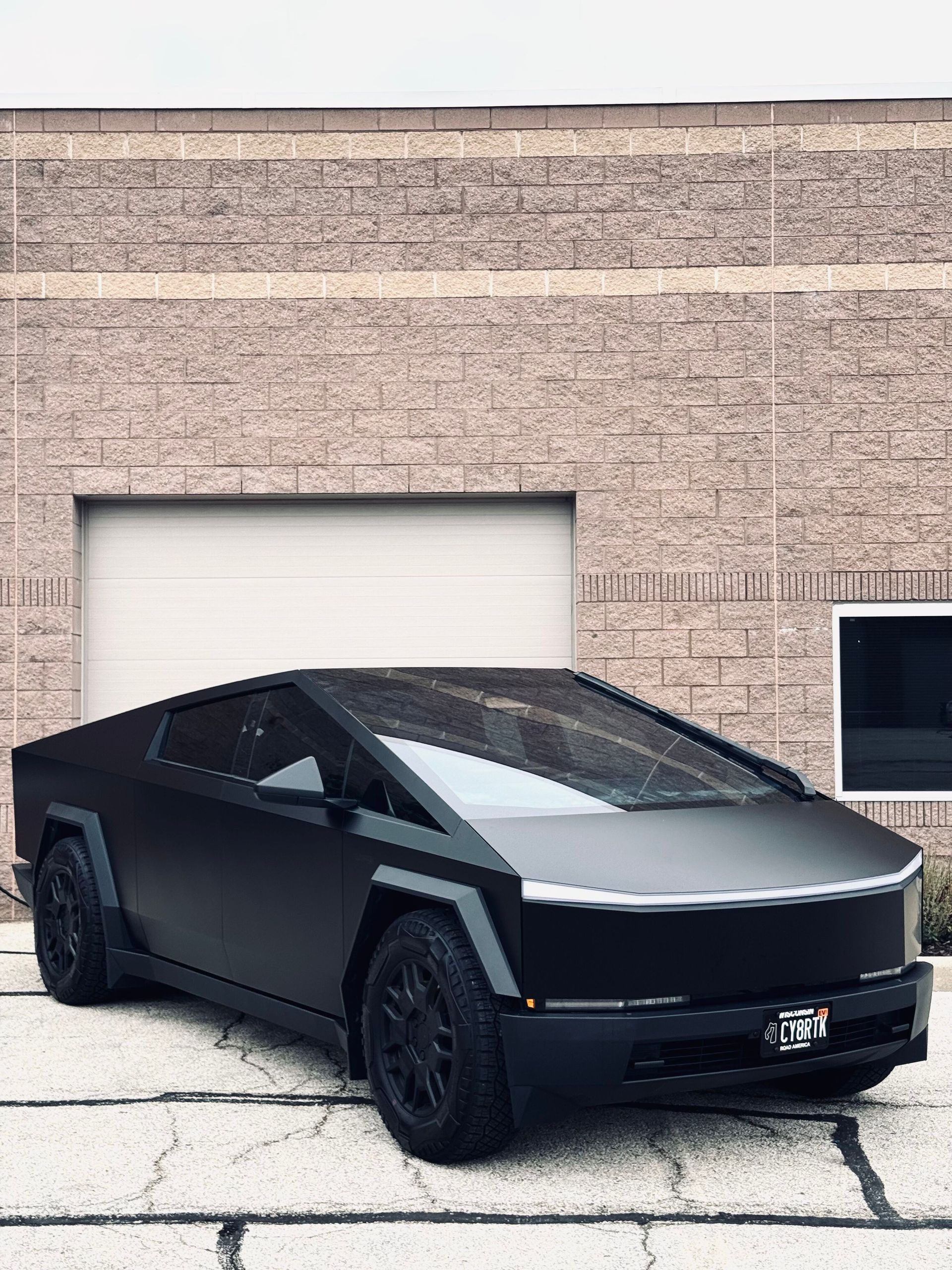 Matte black Tesla Cybertruck parked in front of a brick building with a garage door.