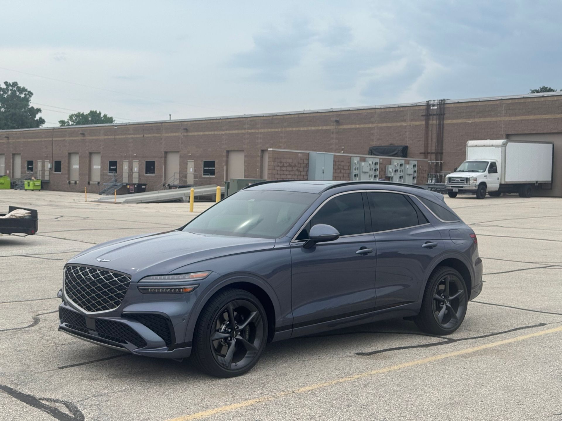 Gray Genesis GV70 SUV parked in front of a brick industrial building. Cloudy sky.