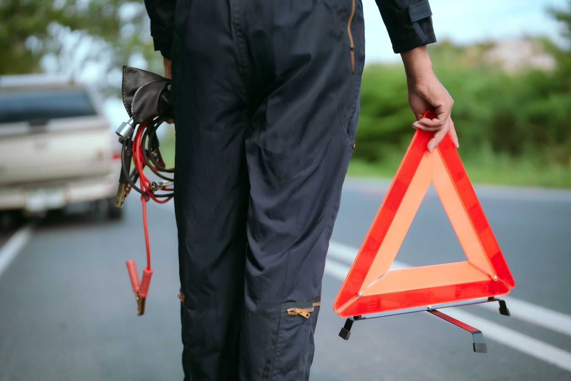 A person in dark clothing stands on a road near a vehicle, holding red jumper cables and a reflective hazard triangle.