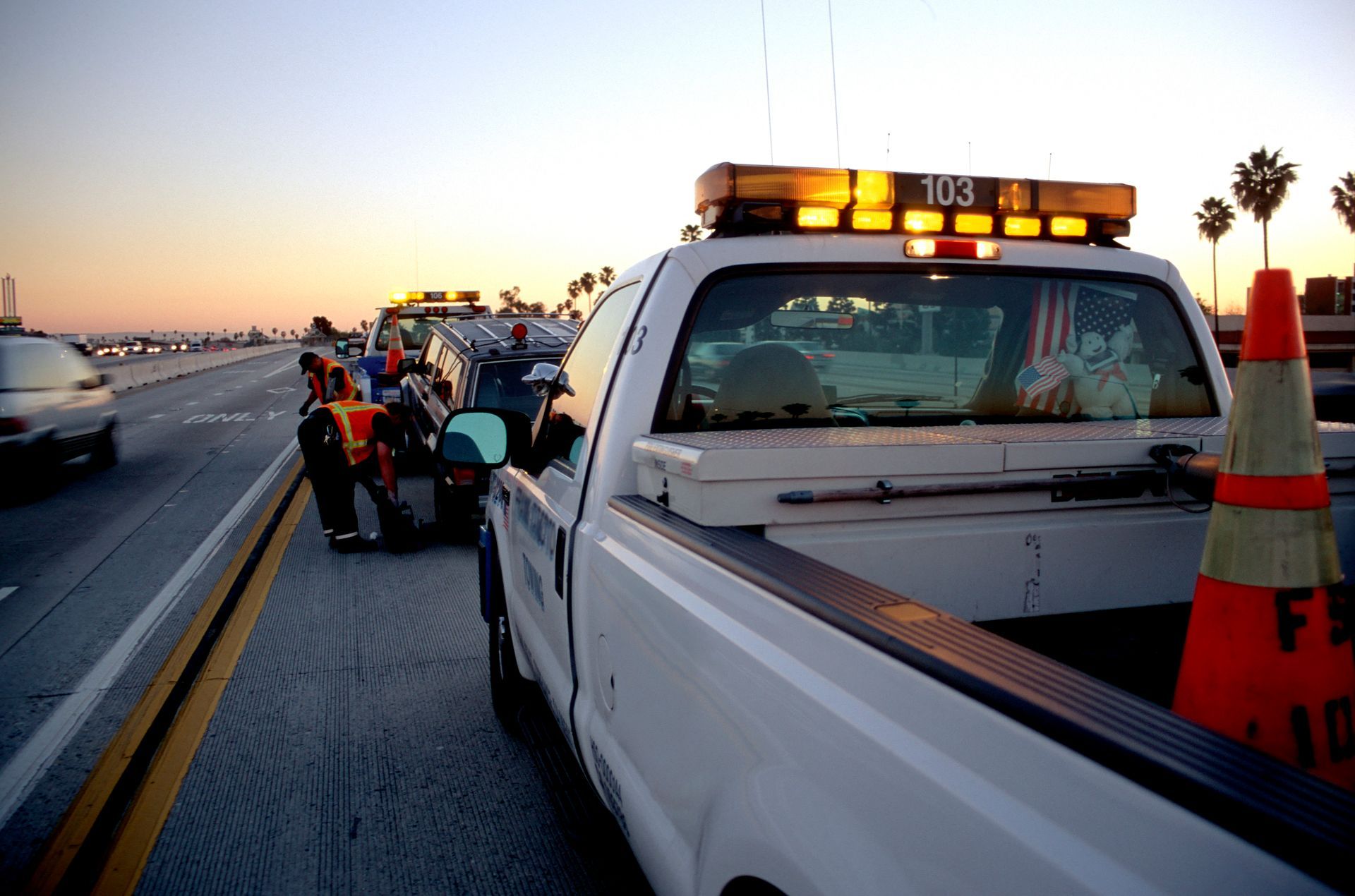 Service truck parked on a highway shoulder at sunset, with a traffic cone and road workers in the background.