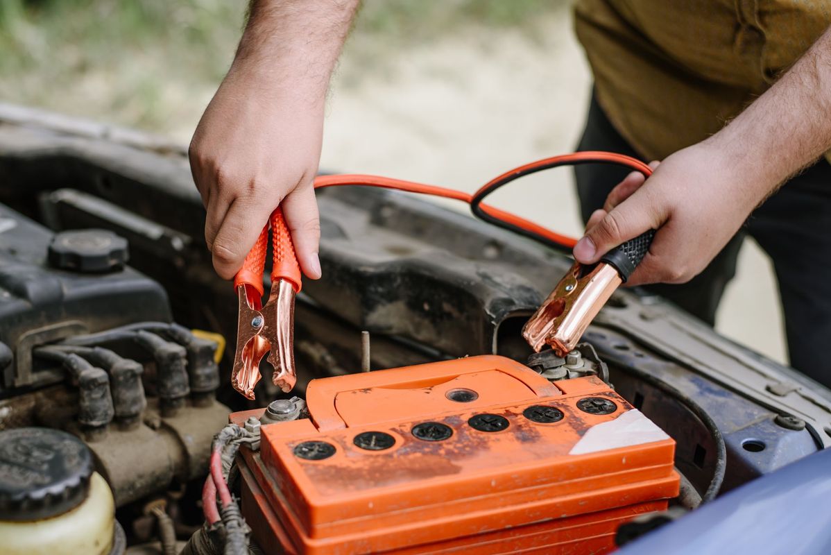 A person holding red and black jumper cable clamps over an orange car battery in an open vehicle hood.