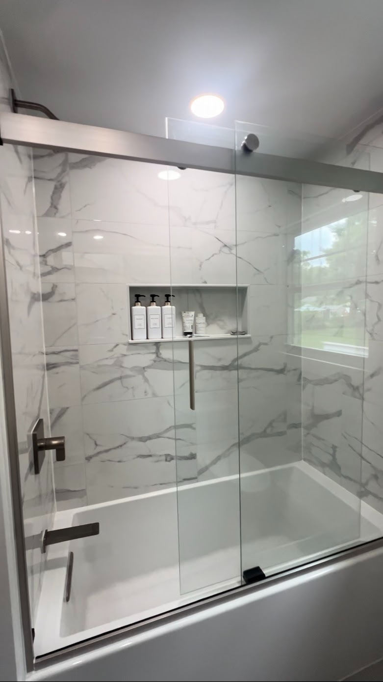 Modern glass shower with white marble-look tile, built-in shelves, and a sliding door in a bright bathroom