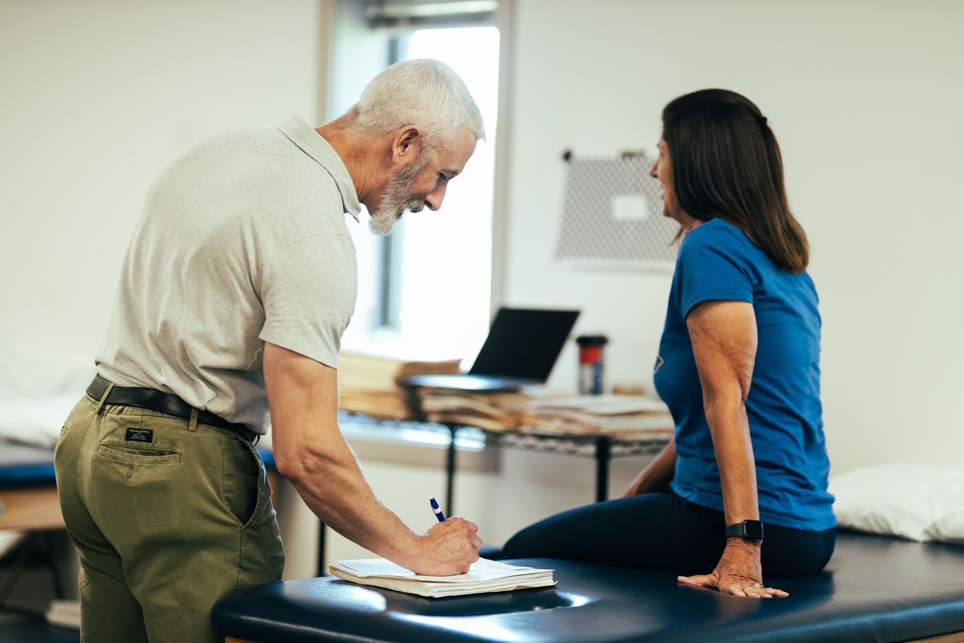 Therapist writing on a clipboard, talking to a woman sitting on a medical table in an exam room.