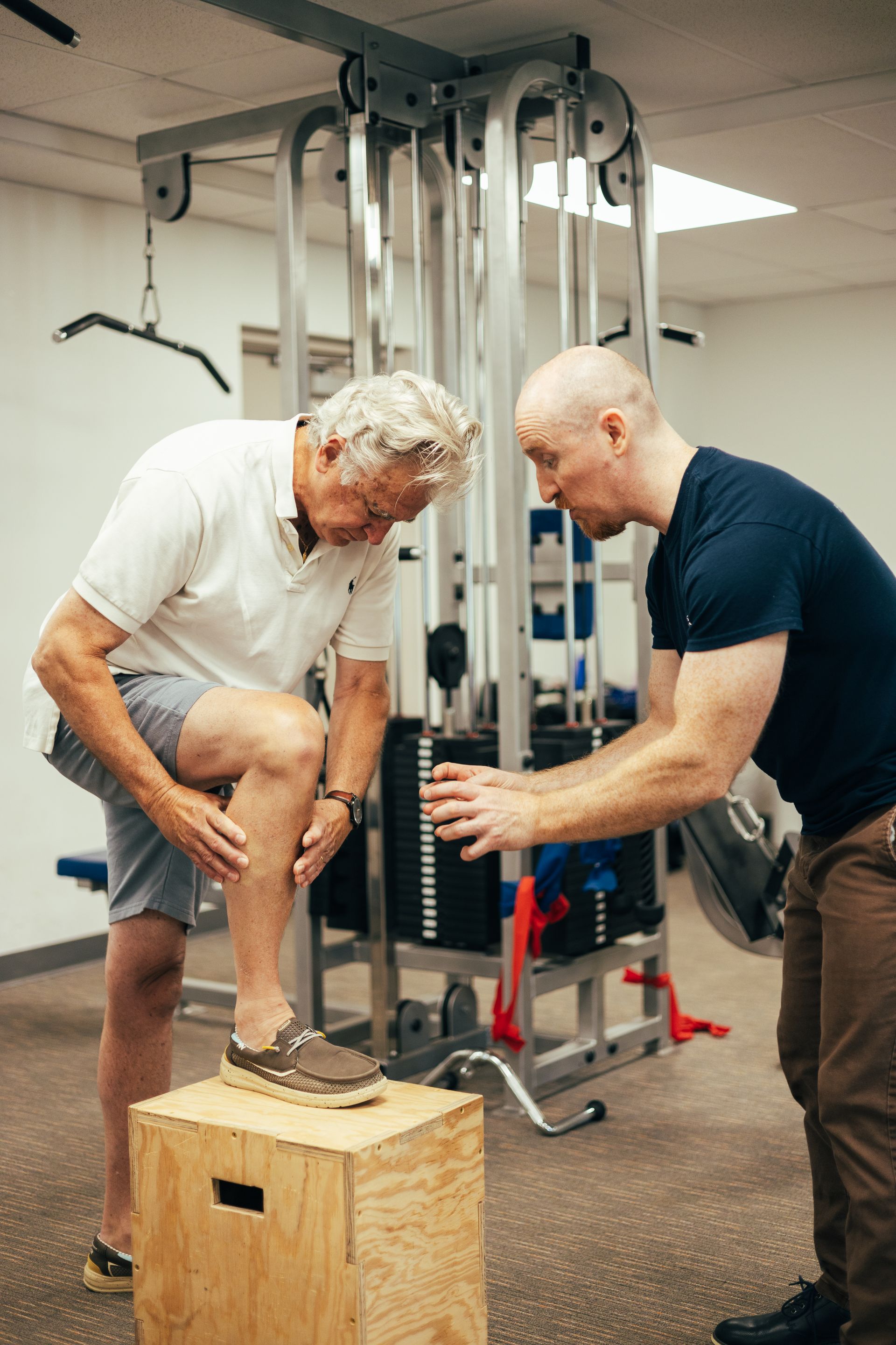 A man receiving physical therapy from an instructor. Man on a box, looking at his leg, in a gym.