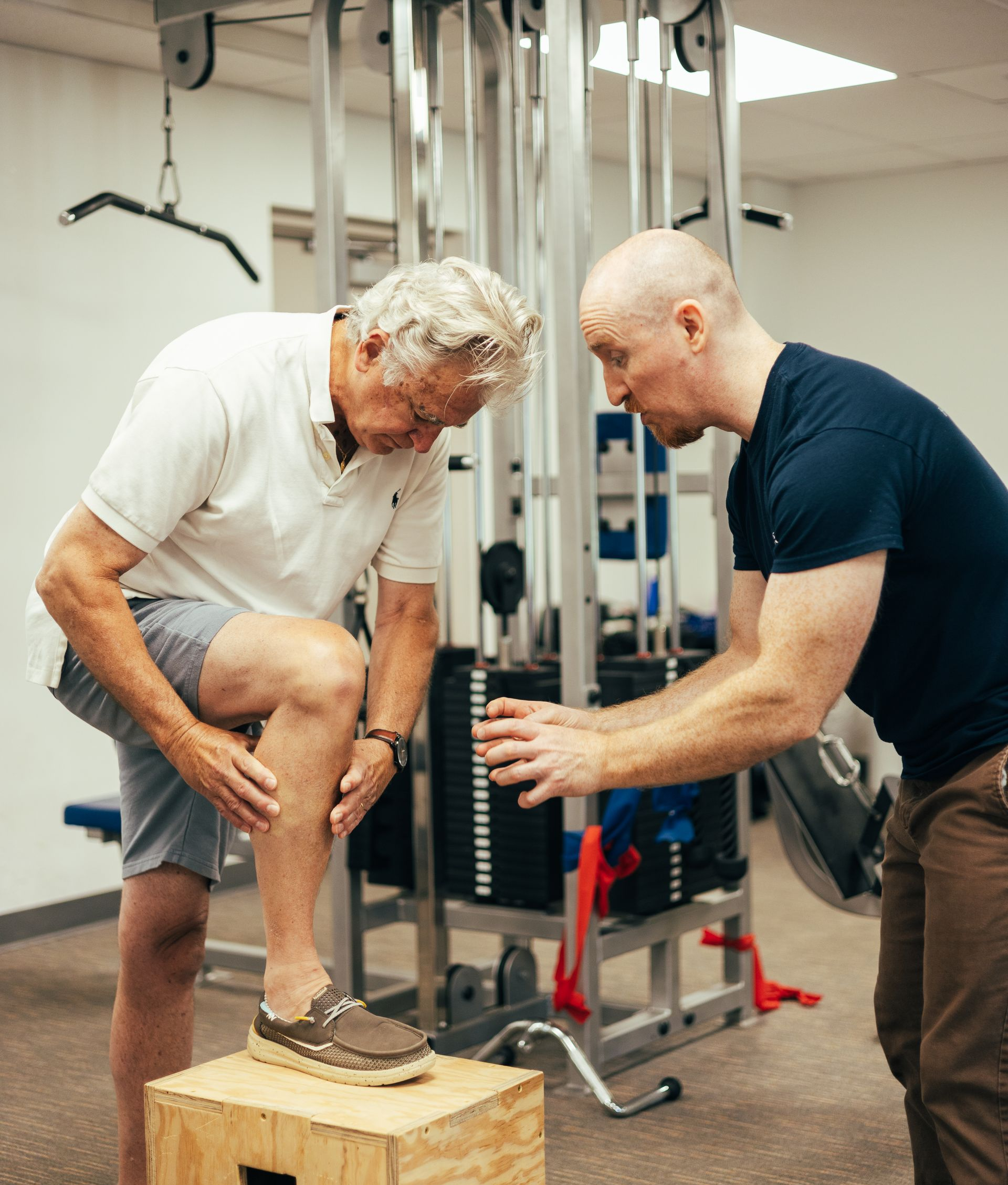 Older man receiving a physical exam from a younger man in a gym. The older man points at his knee.