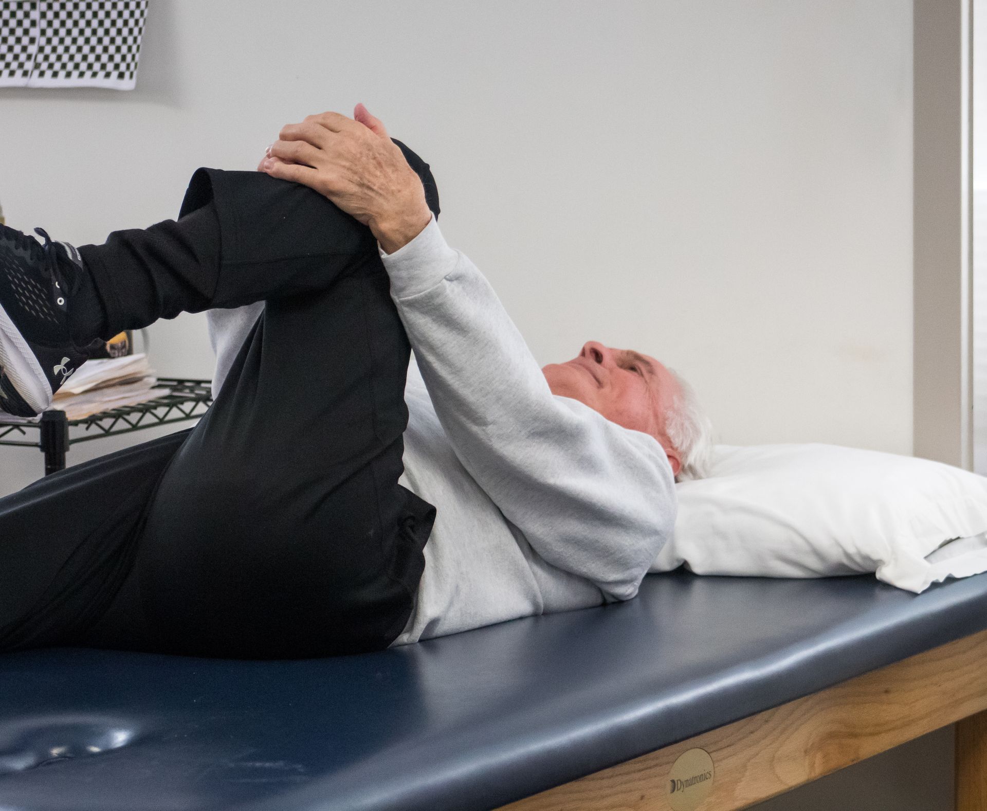 Man on a therapy table, holding his knee to his chest, likely stretching. White walls, black pants, and a white pillow.