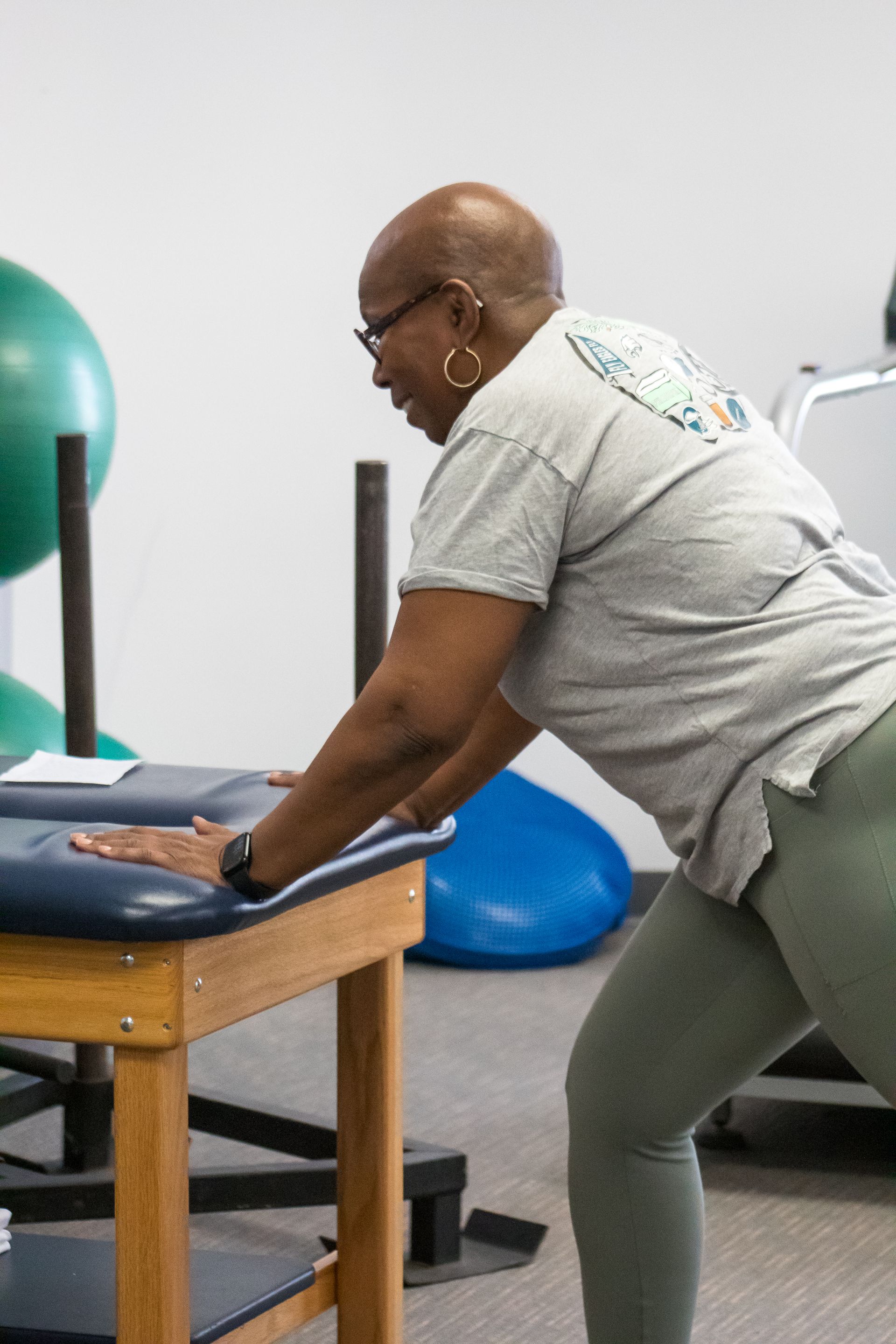 Woman in exercise studio leans on a table, stretching her leg. Blue and green exercise balls visible.