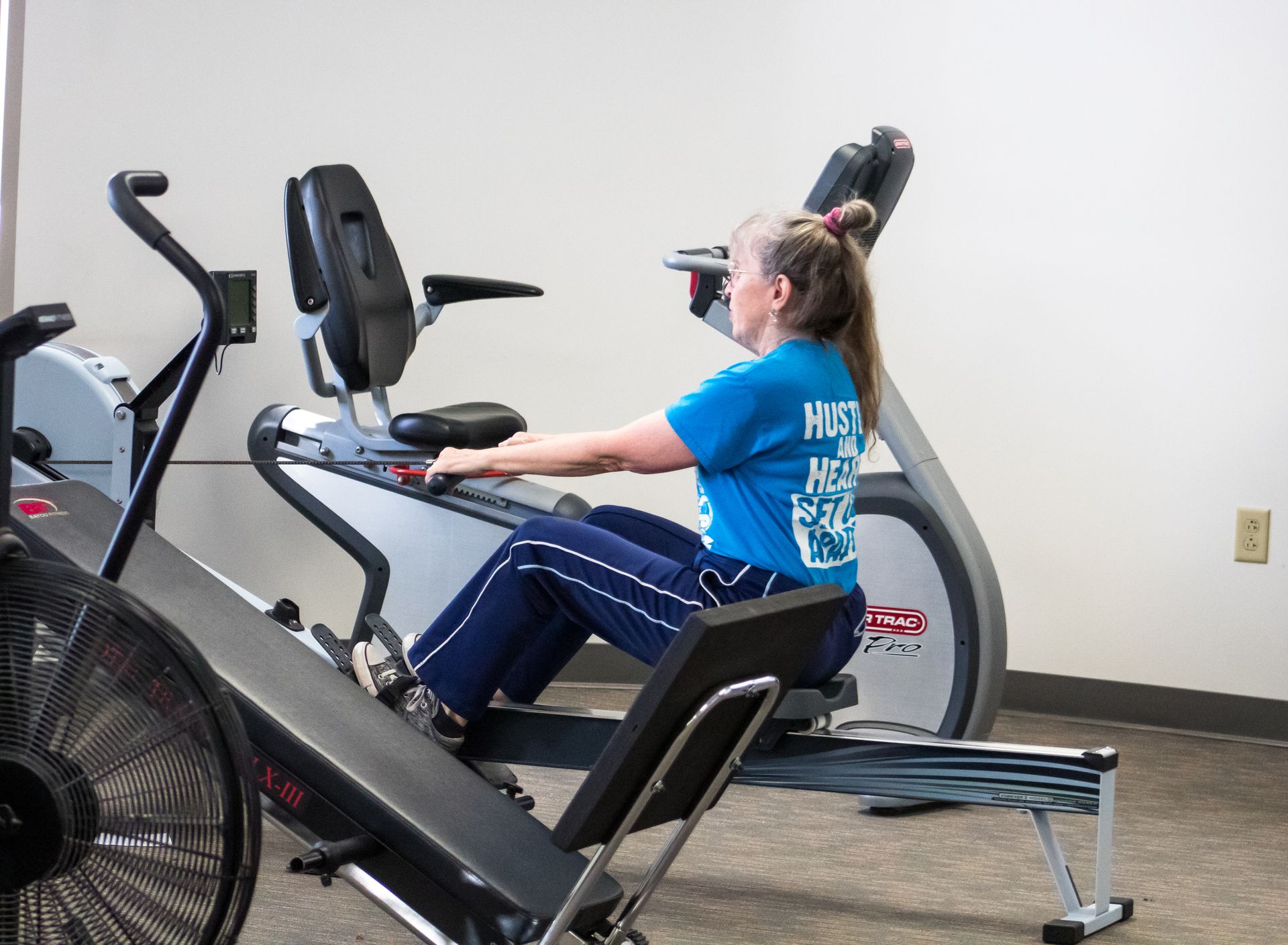 Woman using a rowing machine in a gym setting. She wears a blue shirt and dark pants.