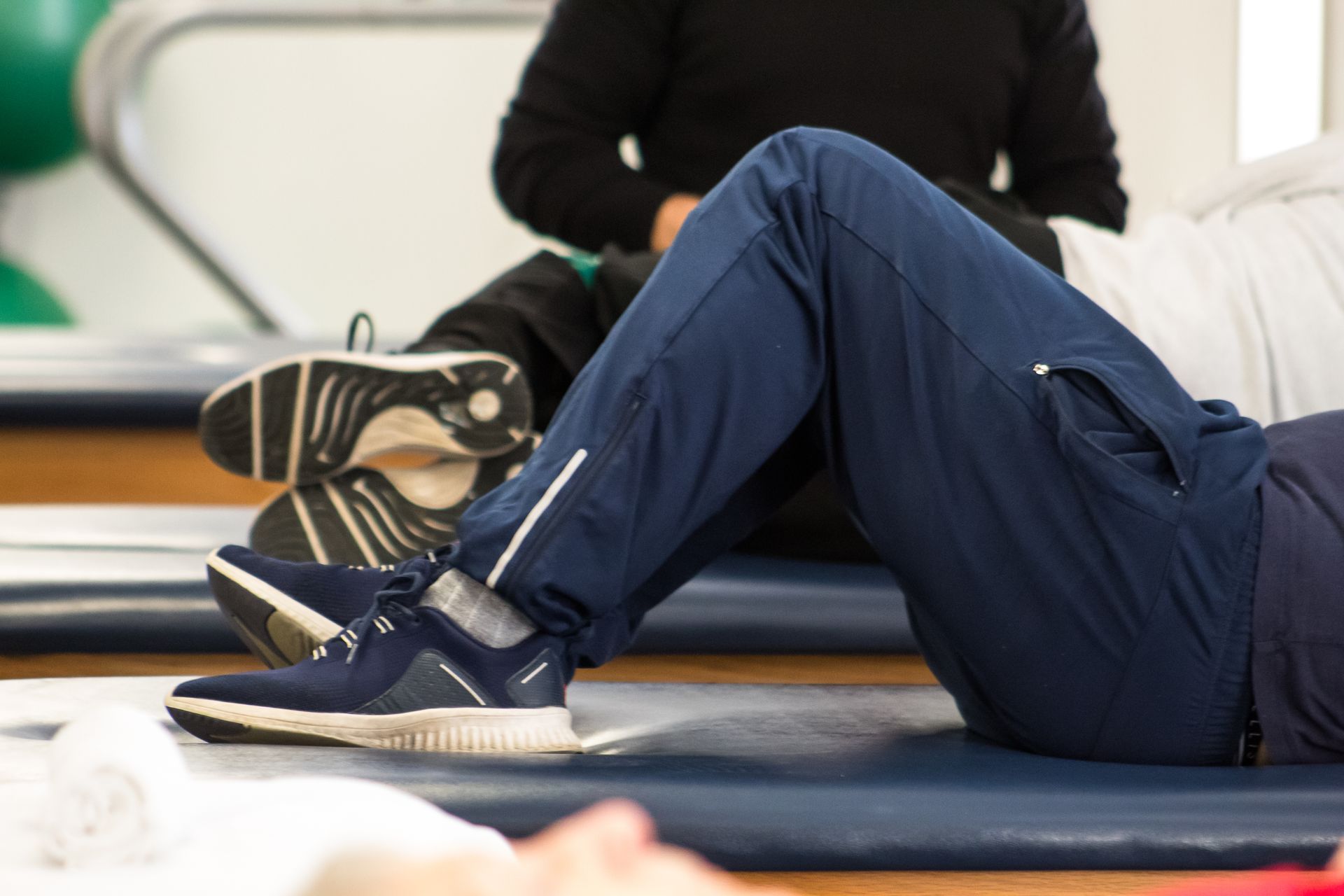 Person in blue athletic pants and shoes, performing exercise on a mat, with a second person seated nearby.