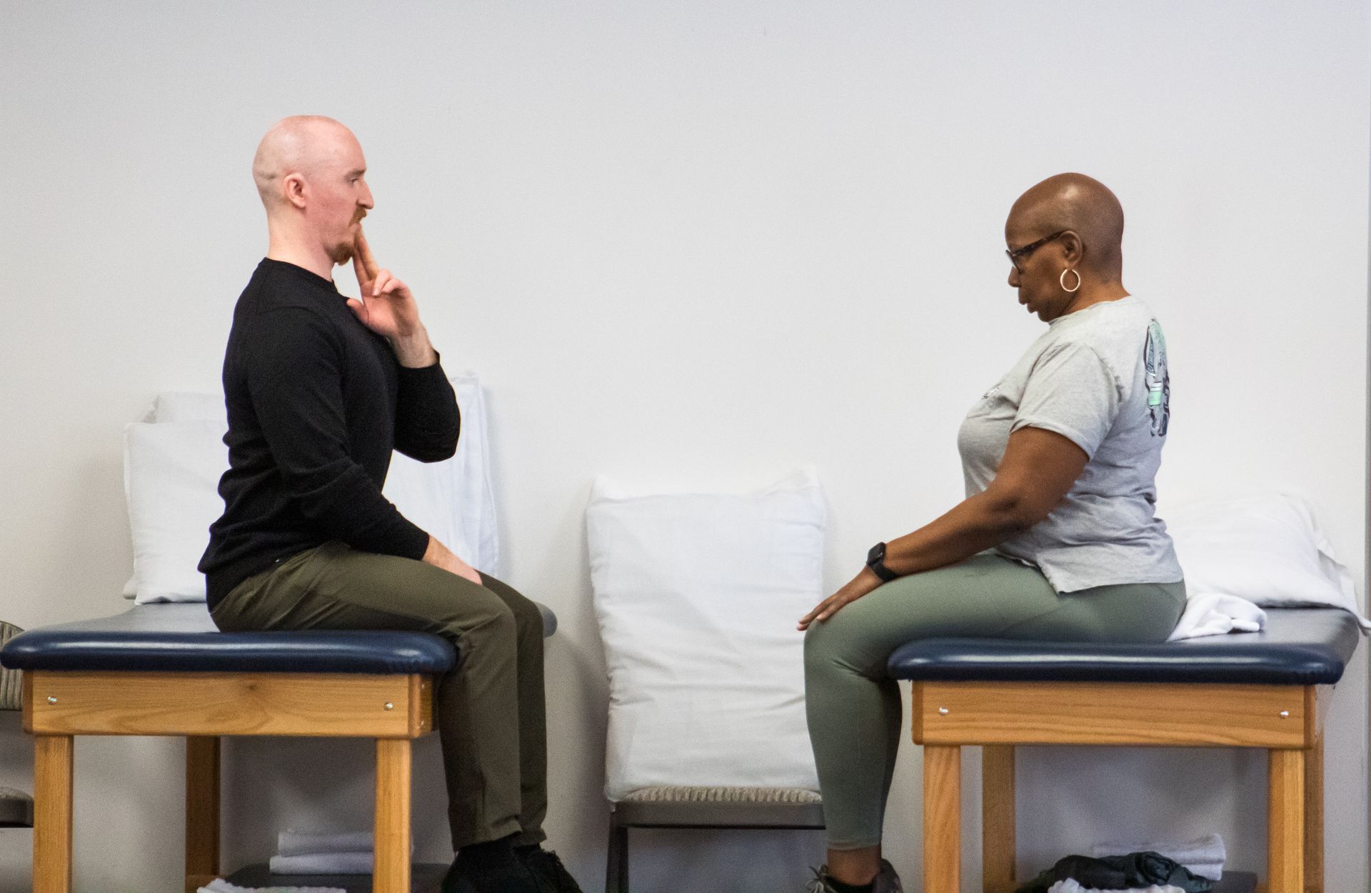 Man and woman seated, facing each other. Man holds fingers near his face. Both sit on therapy tables in a white room.