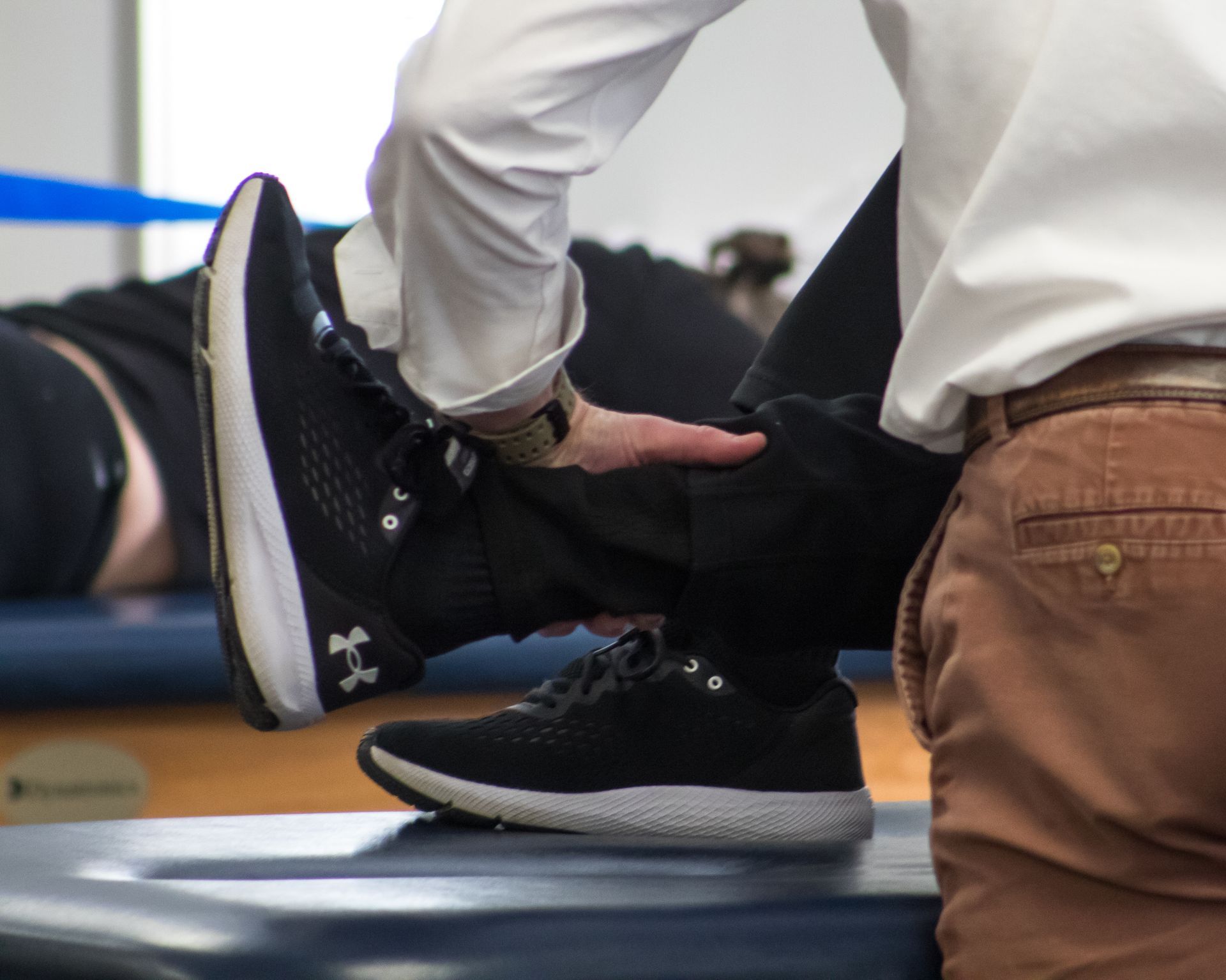 A therapist stretches a patient's leg. Person in white shirt and tan pants; patient on a blue table.