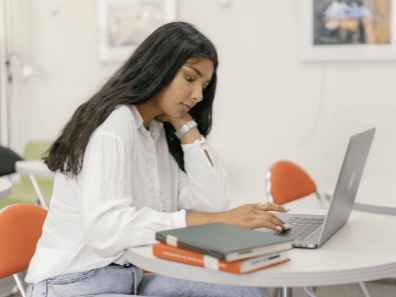 Woman with long dark hair using laptop at a table, books in front of her. Bright room, focus on the screen.