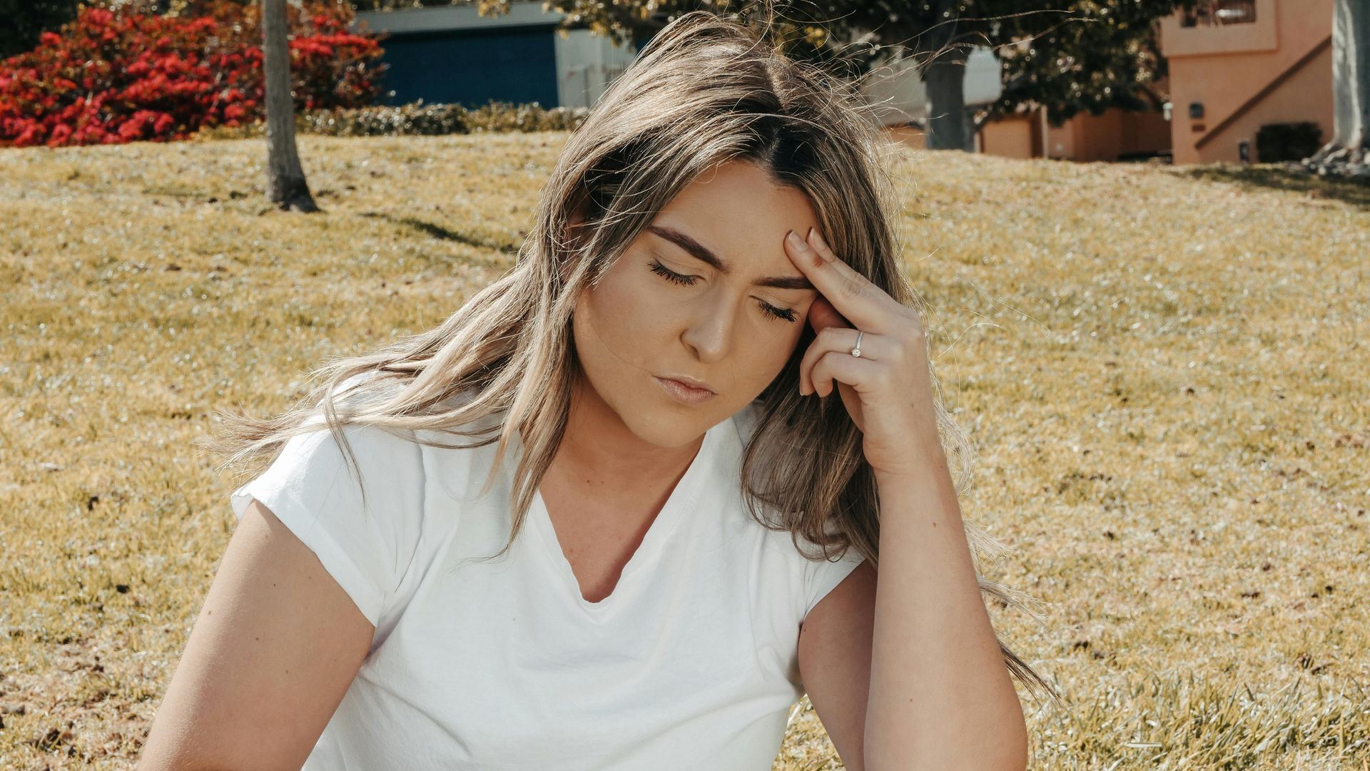 A woman is sitting in the grass with her hand on her forehead.