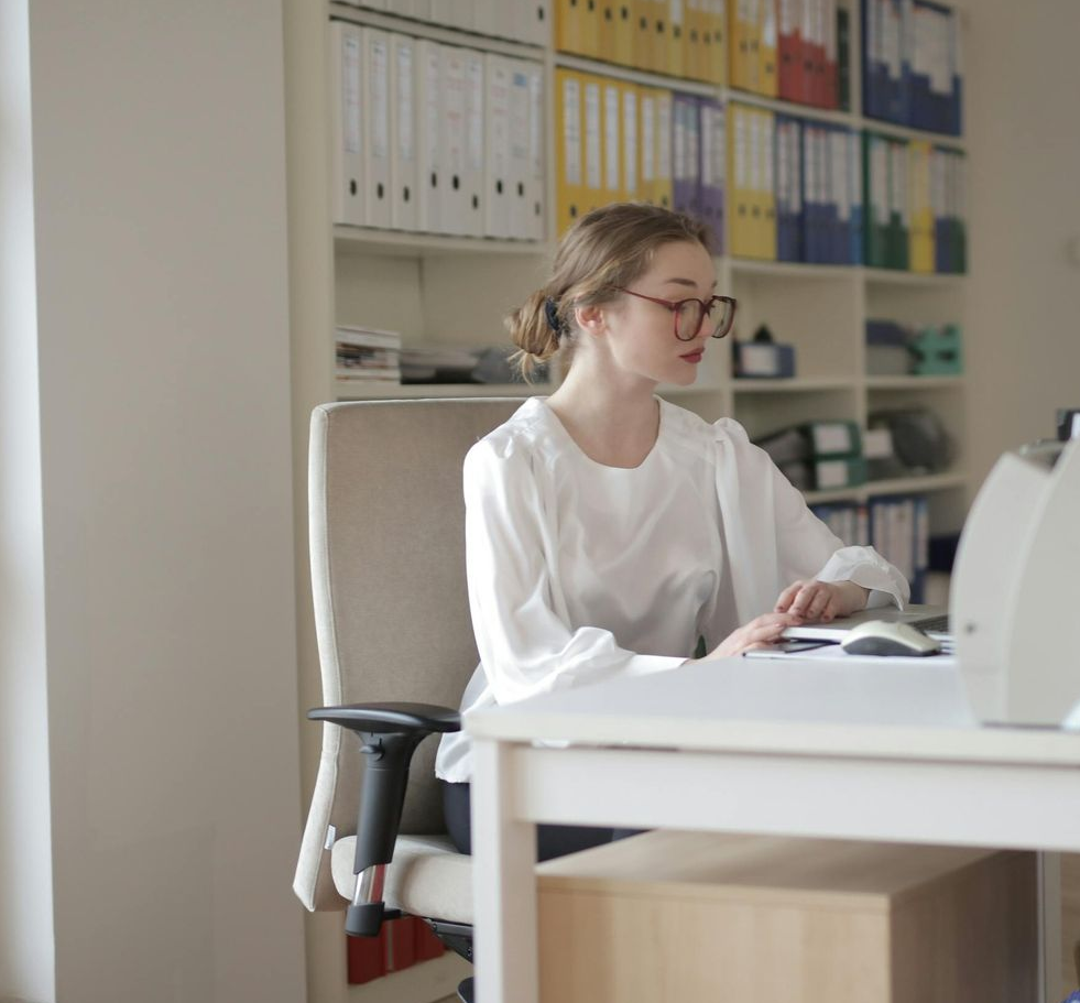 A person wearing glasses and a white blouse sits at a white desk in an office, reading a book in front of shelf binders.