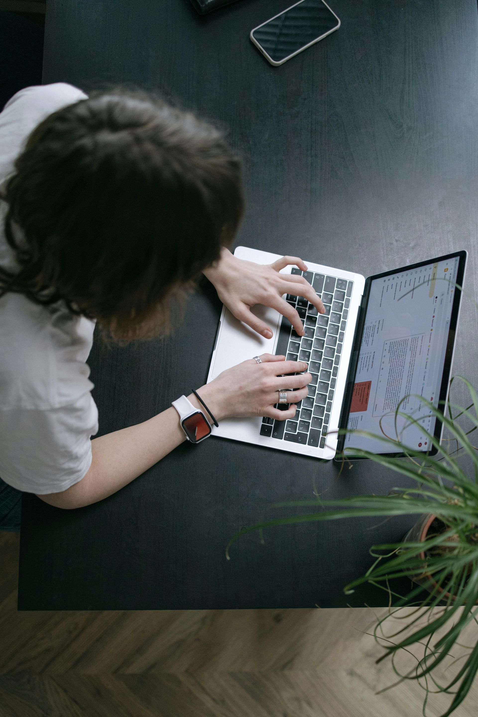 A person typing on a laptop at a dark desk next to a potted plant, captured from an overhead, high-angle view.