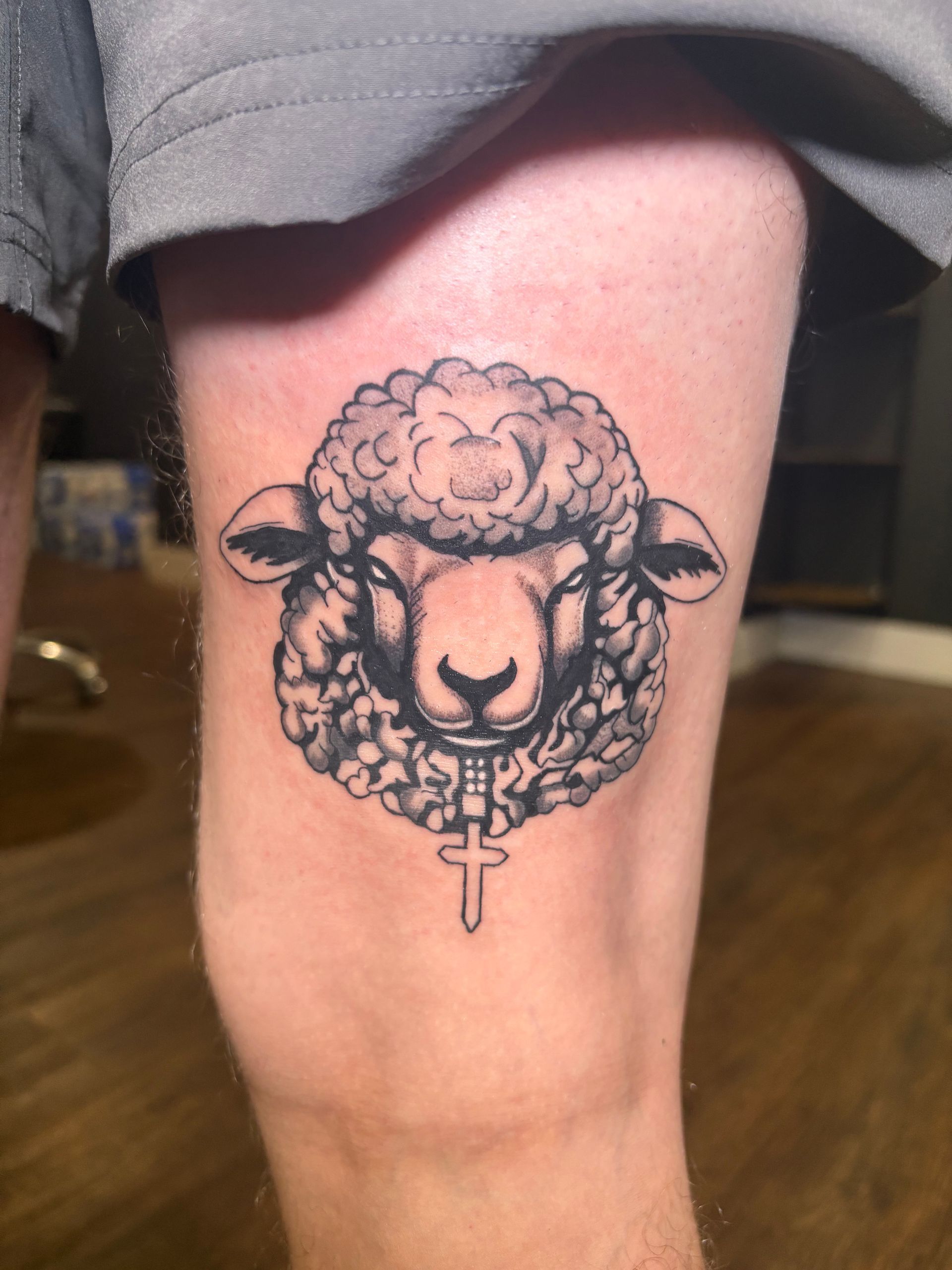 Black and white tattoo of a sheep head with a cross pendant on a person's knee.