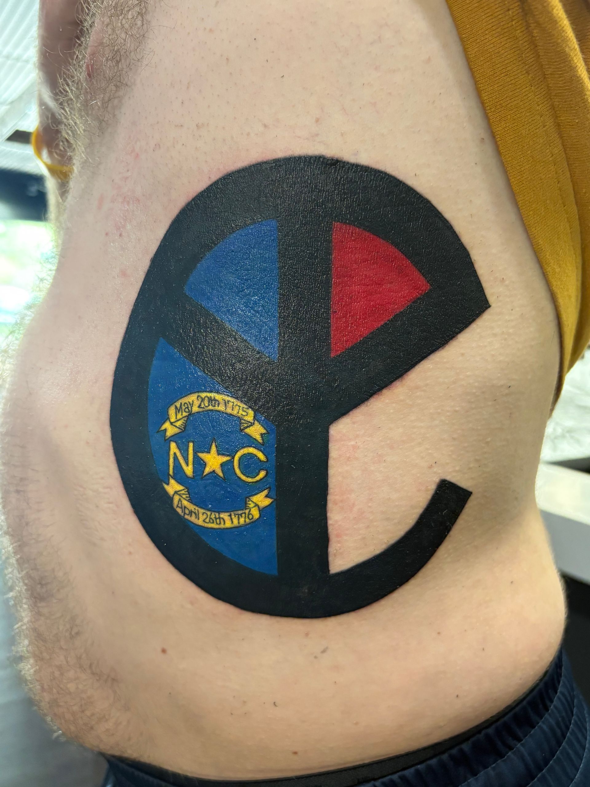 Tattoo of the North Carolina state flag symbol inside a peace sign on a person's side, featuring blue, red, and black.