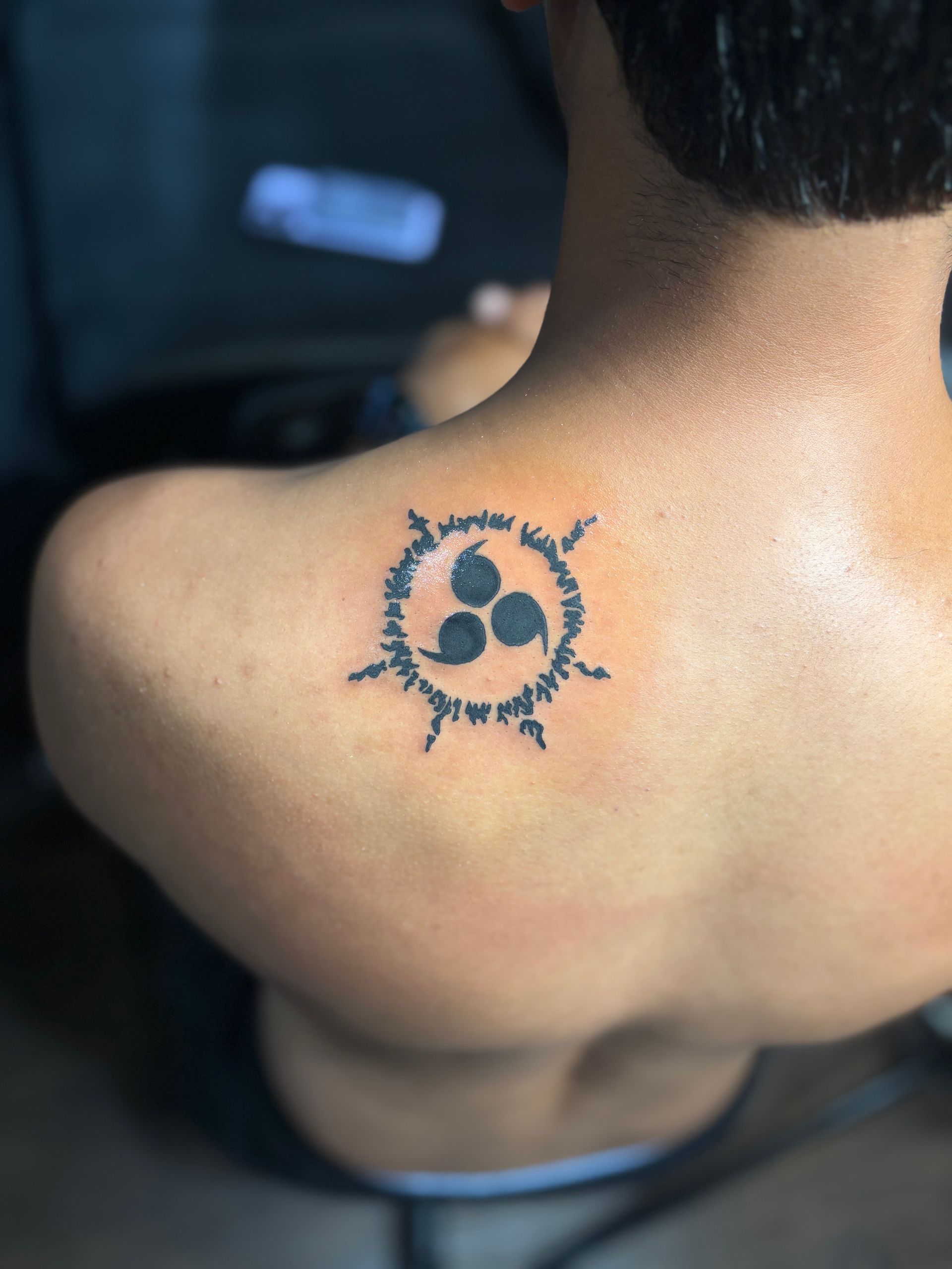 Tattoo on the upper back depicting a black, spiral-like design with three comma shapes in the center.