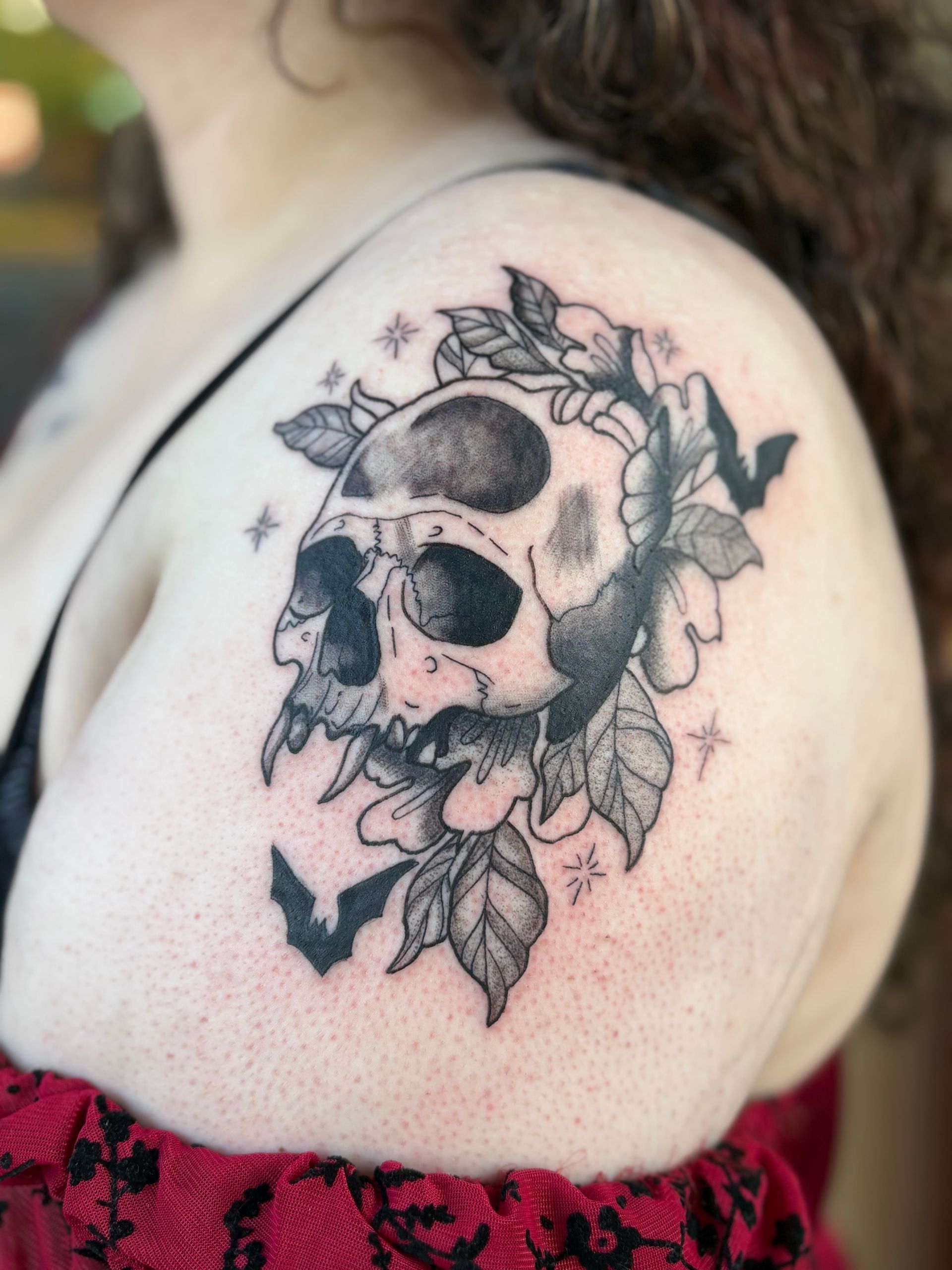 Tattoo of a skull with flowers and bats on a person's shoulder, in black ink with stars.